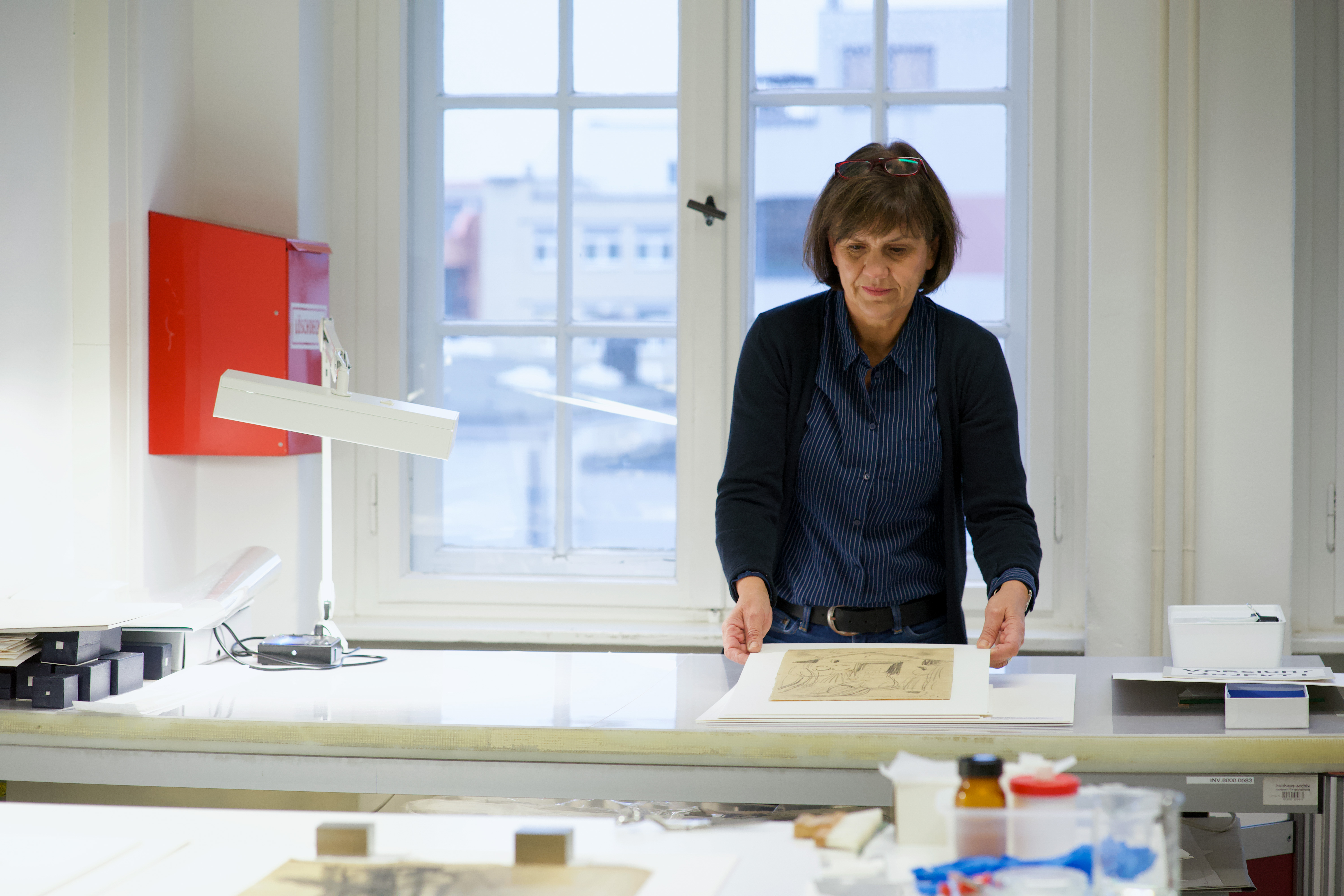 Eine Frau schaut sich Kunstwerke an einem Tisch in einem hellen Raum mit großen Fenstern an.