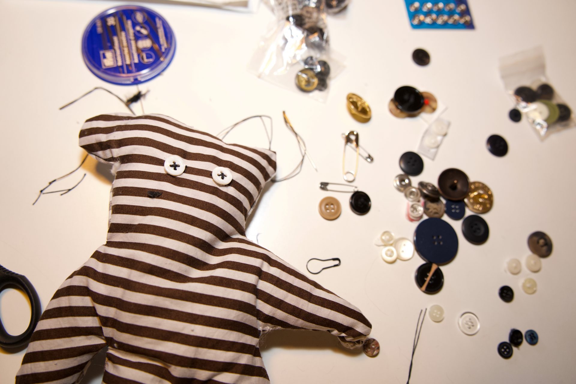Gestreifter Stoffbär mit Knöpfen und Nähmaterial, umgeben von verschiedenen Knöpfen und Nähutensilien.