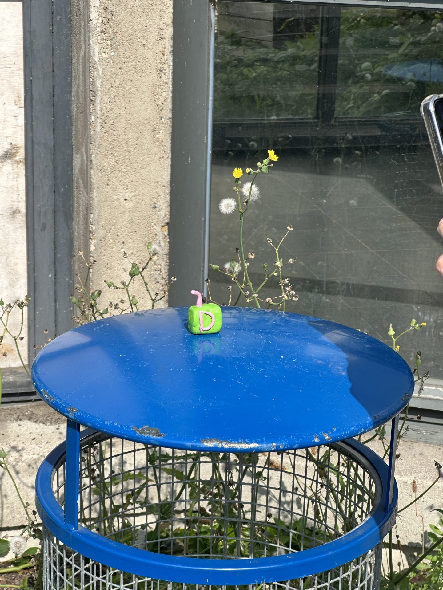 Blaue Mülltonne mit buntem Dekoobjekt, umgeben von grünen Pflanzen und Fenstern im Hintergrund.