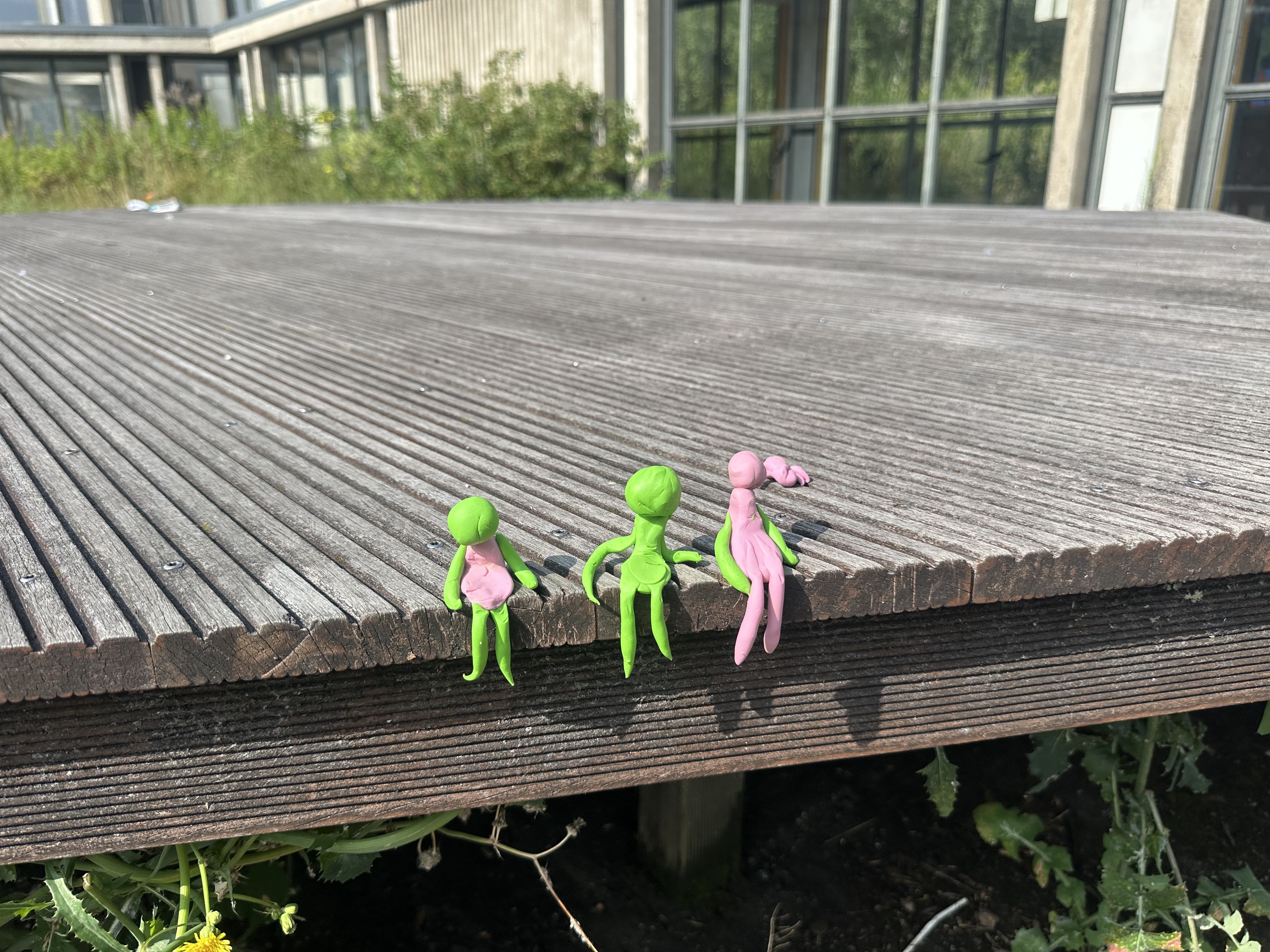 Drei kleine, bunte Figuren sitzen auf einer Holzoberfläche, umgeben von Pflanzen und einem modernen Gebäude im Hintergrund.