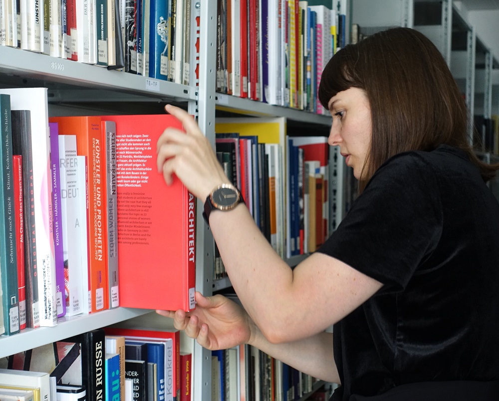 Frau mit braunen Haaren schaut sich Bücher im Regal an und zieht eines heraus.