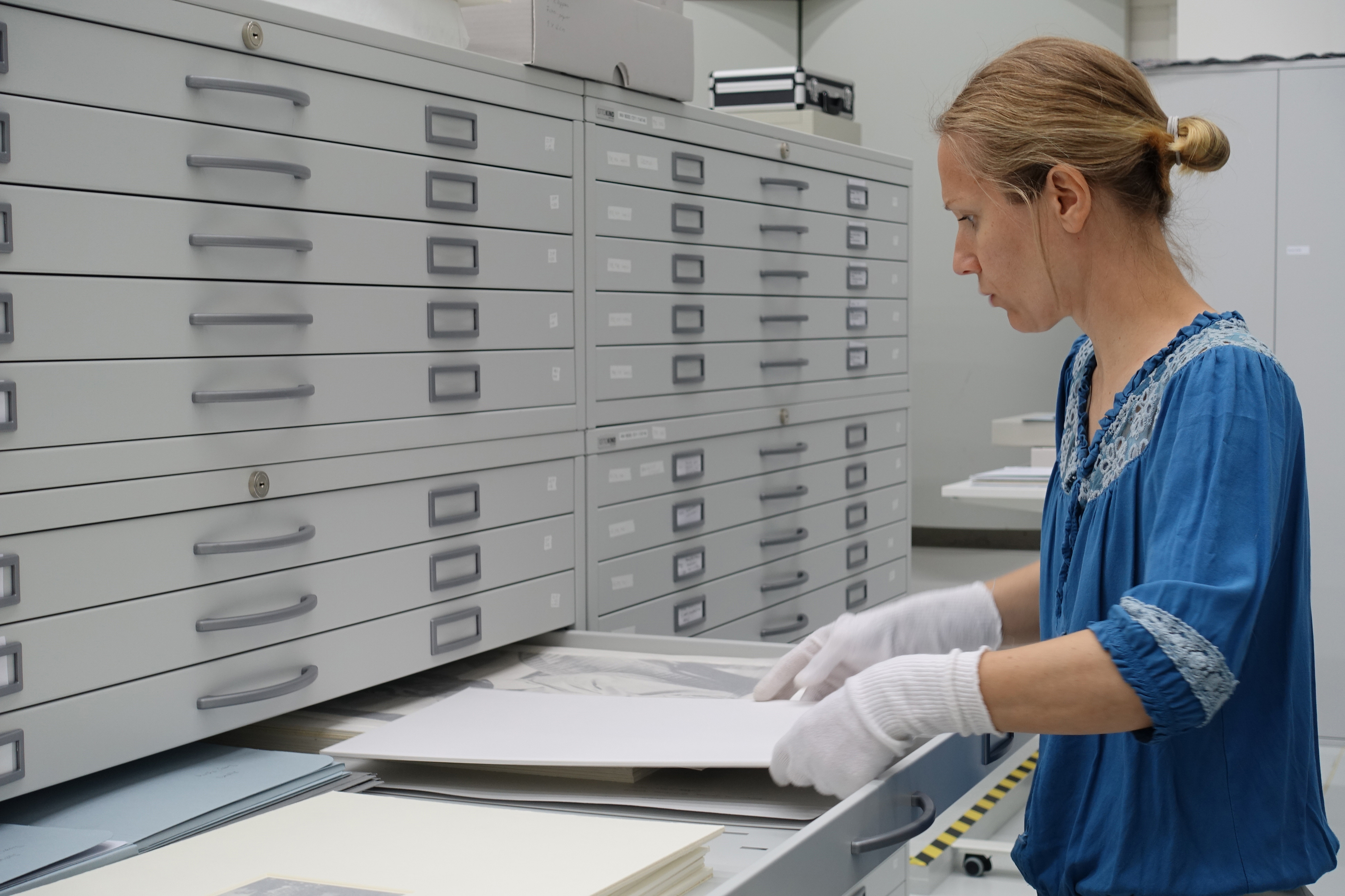 Frau in blauer Bluse sortiert Dokumente in einem Archivschrank. Weiße Handschuhe schützen die Unterlagen.
