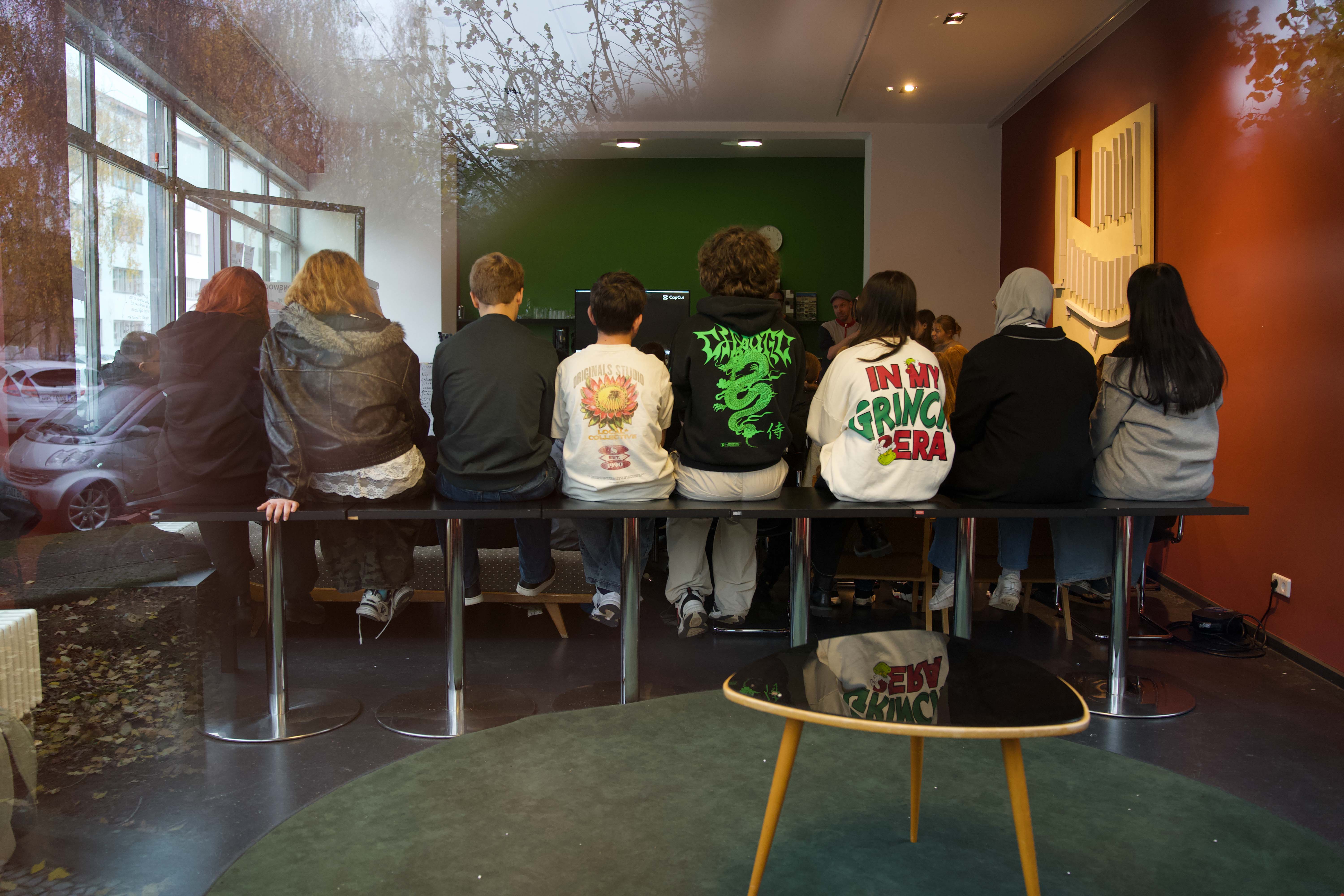Eine Gruppe junger Menschen sitzt auf einer Bank und blickt in einen Raum mit grünen Wänden und Möbeln.