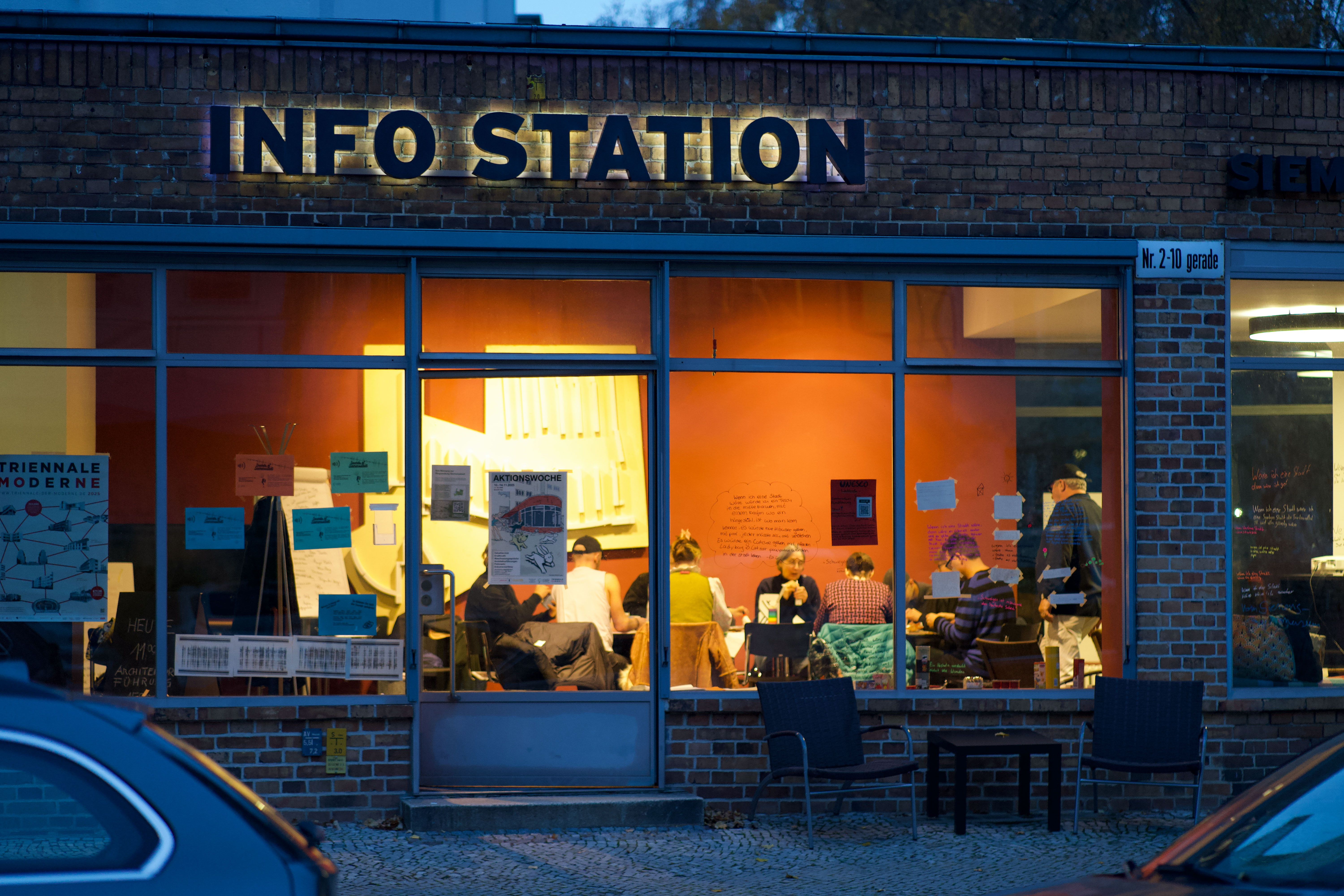 Info-Station: Ein heller Raum mit mehreren Personen, umgeben von bunten Plakaten hinter großen Fenstern.