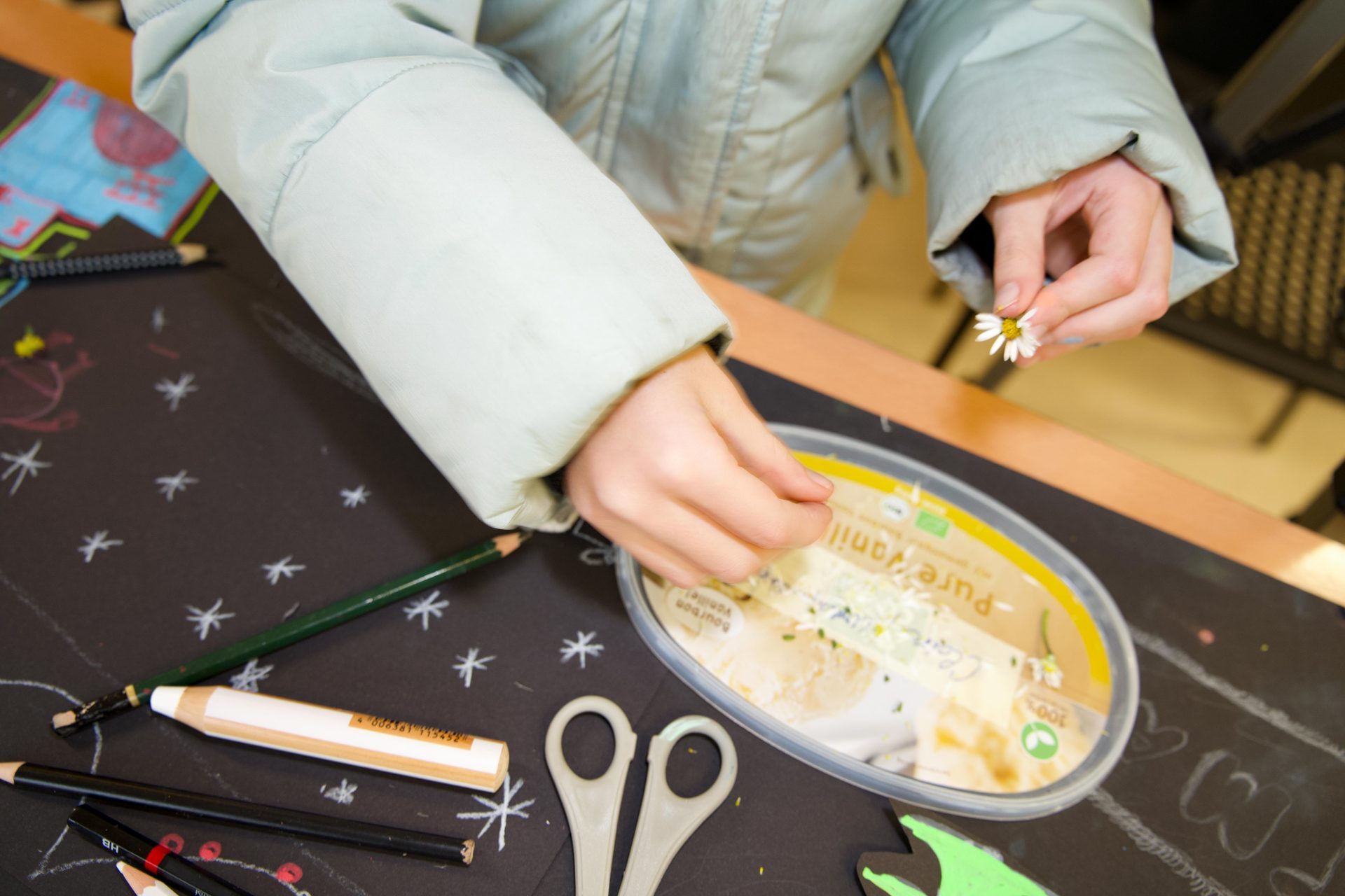 Eine Hand hält eine Blume, während der Tisch mit Stiften und Bastelmaterialien gedeckt ist.