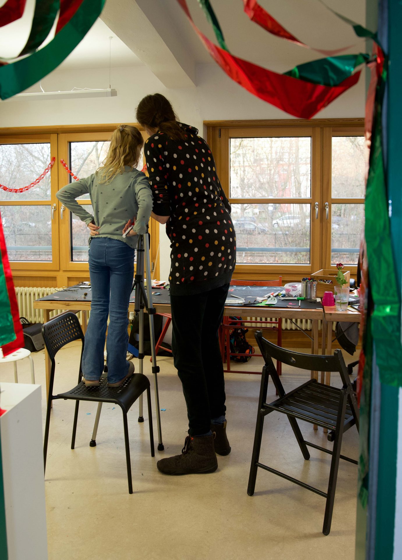 Ein Kind steht auf einem Stuhl, während ein Erwachsener ihm in einem festlich dekorierten Raum hilft.
