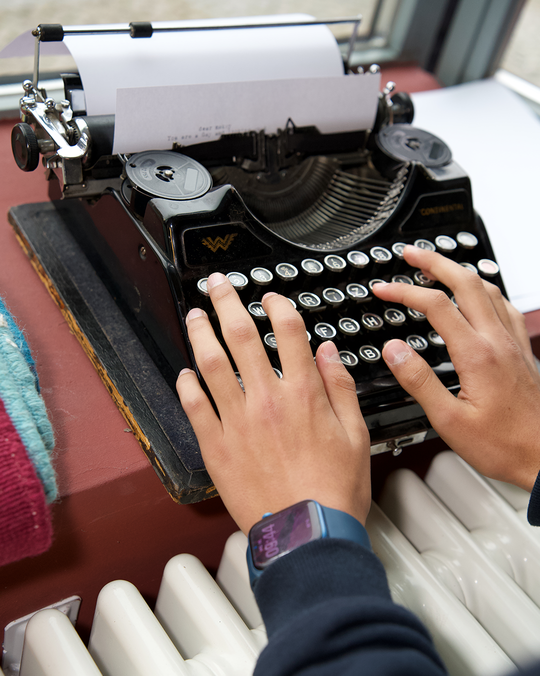 Hands typing on an old typewriter, next to a radiator and a colorful towel.