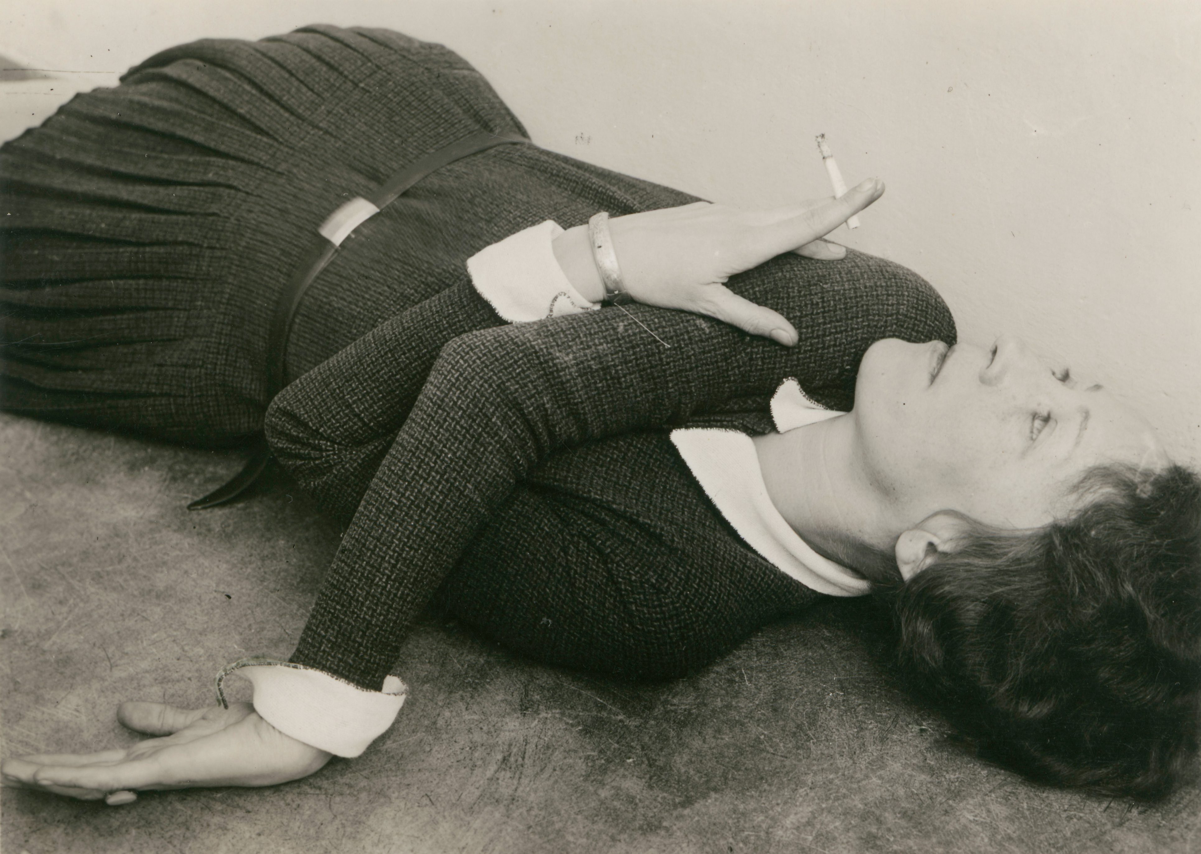 A woman in an elegant outfit lies on the floor smoking a cigarette, looking up at the ceiling.