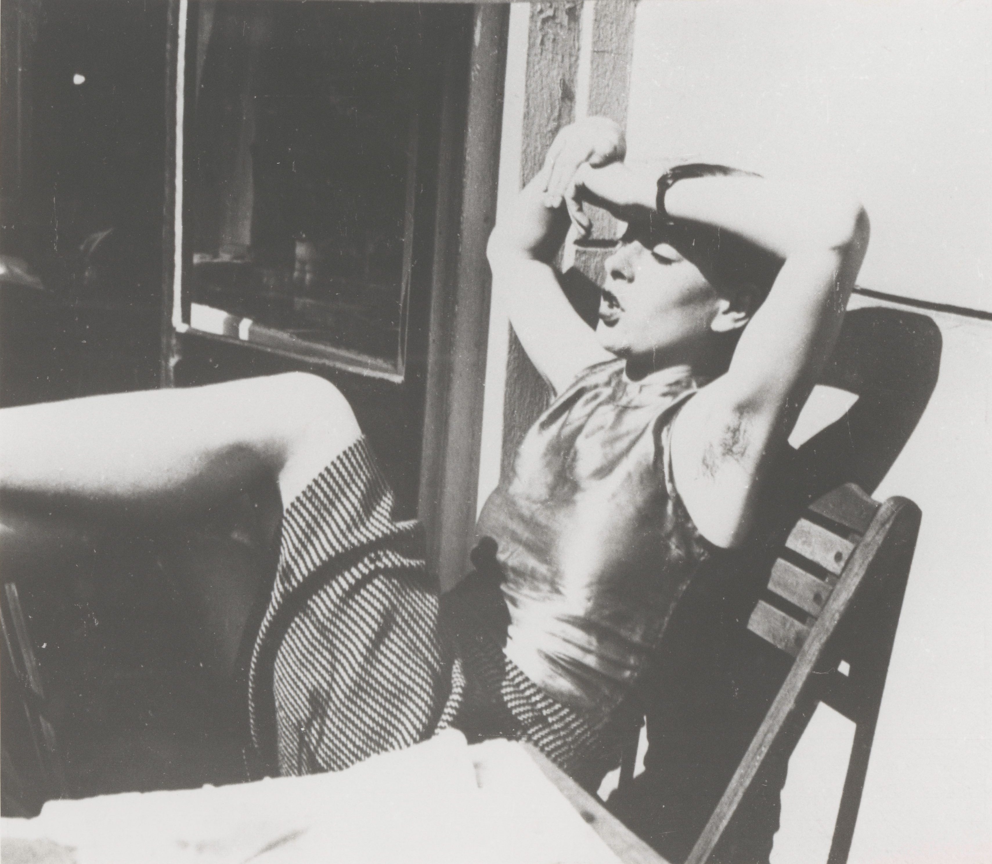 A woman in a striped dress sits relaxed with her arms raised in a chair by a window.