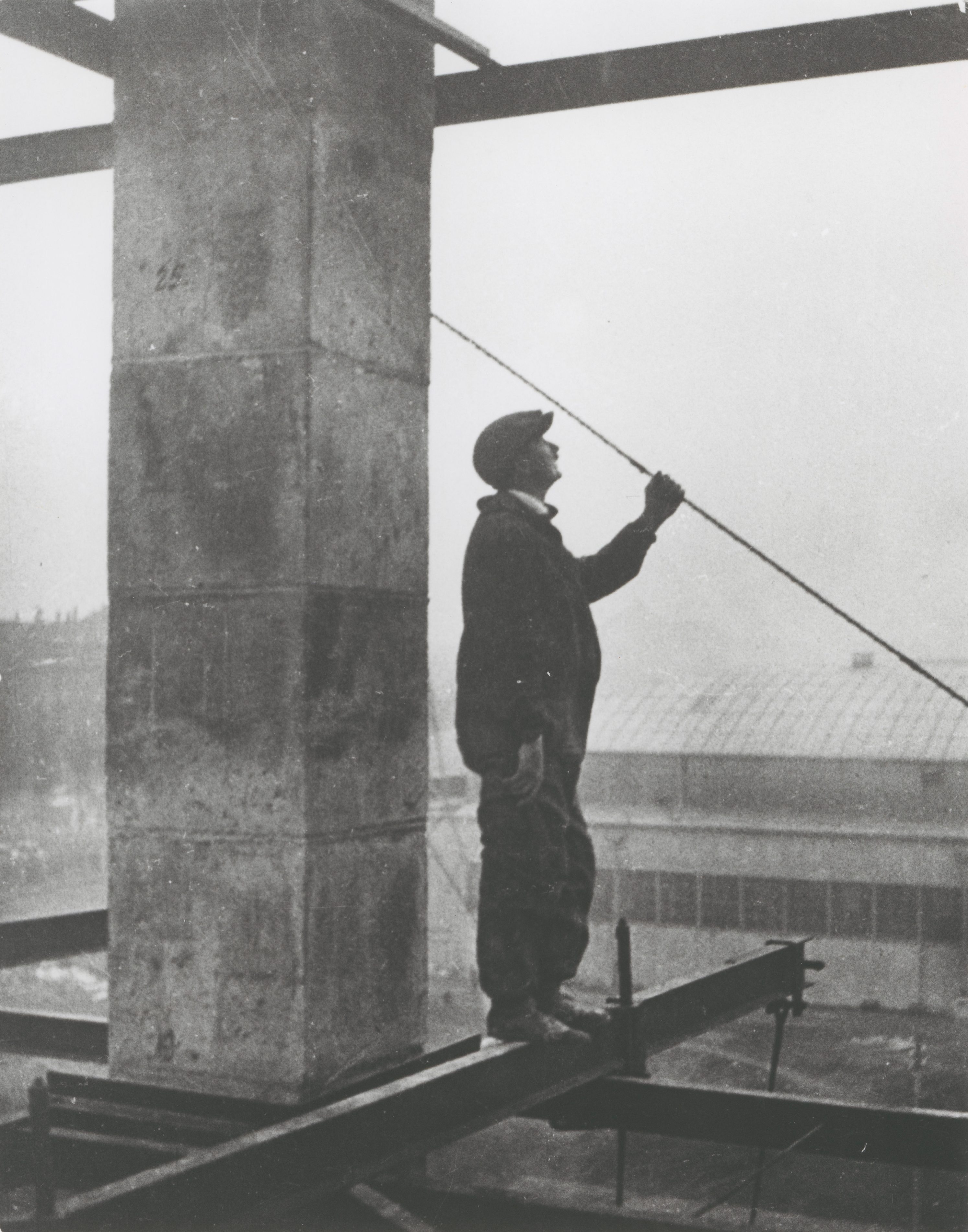 Ein Arbeiter steht auf einer Stahltraverse, hält ein Seil, im Hintergrund ein unfertiges Gebäude und eine neblige Atmosphäre.