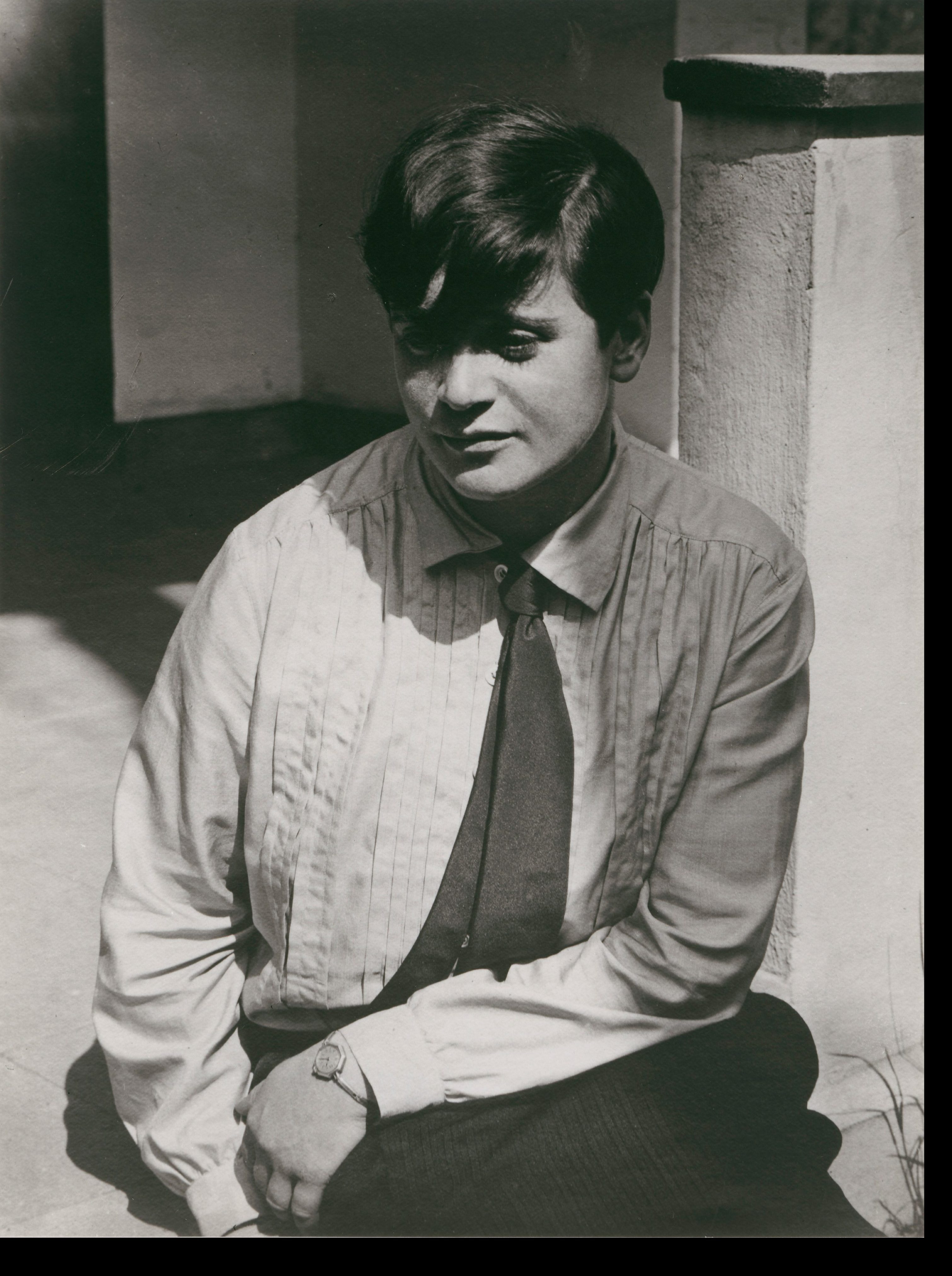 A young man sits outdoors, wearing a light shirt and tie, with a serious expression on his face.