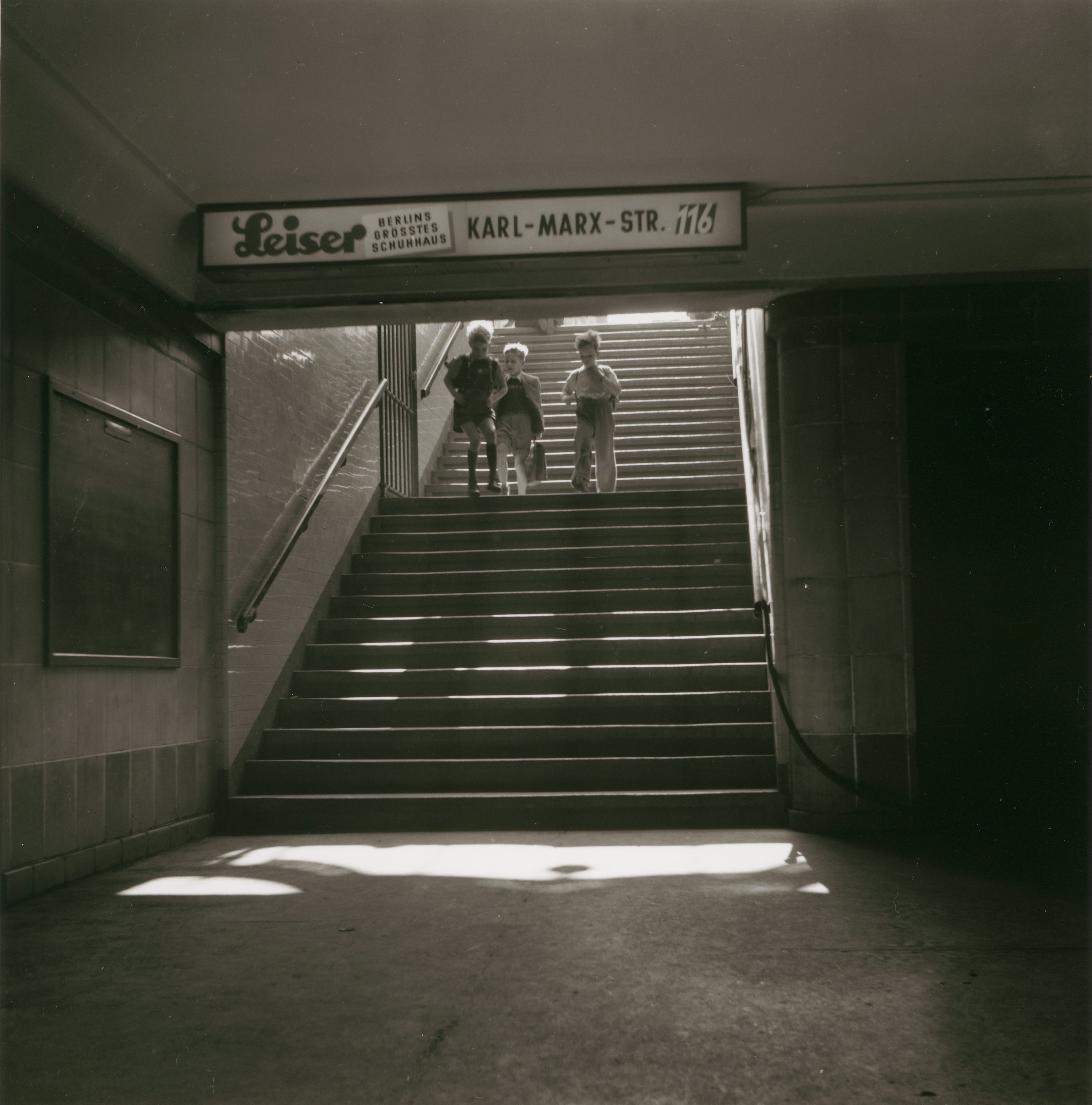 Black-and-white image of children going up stairs, with a neon sign for "Leiser" above them.