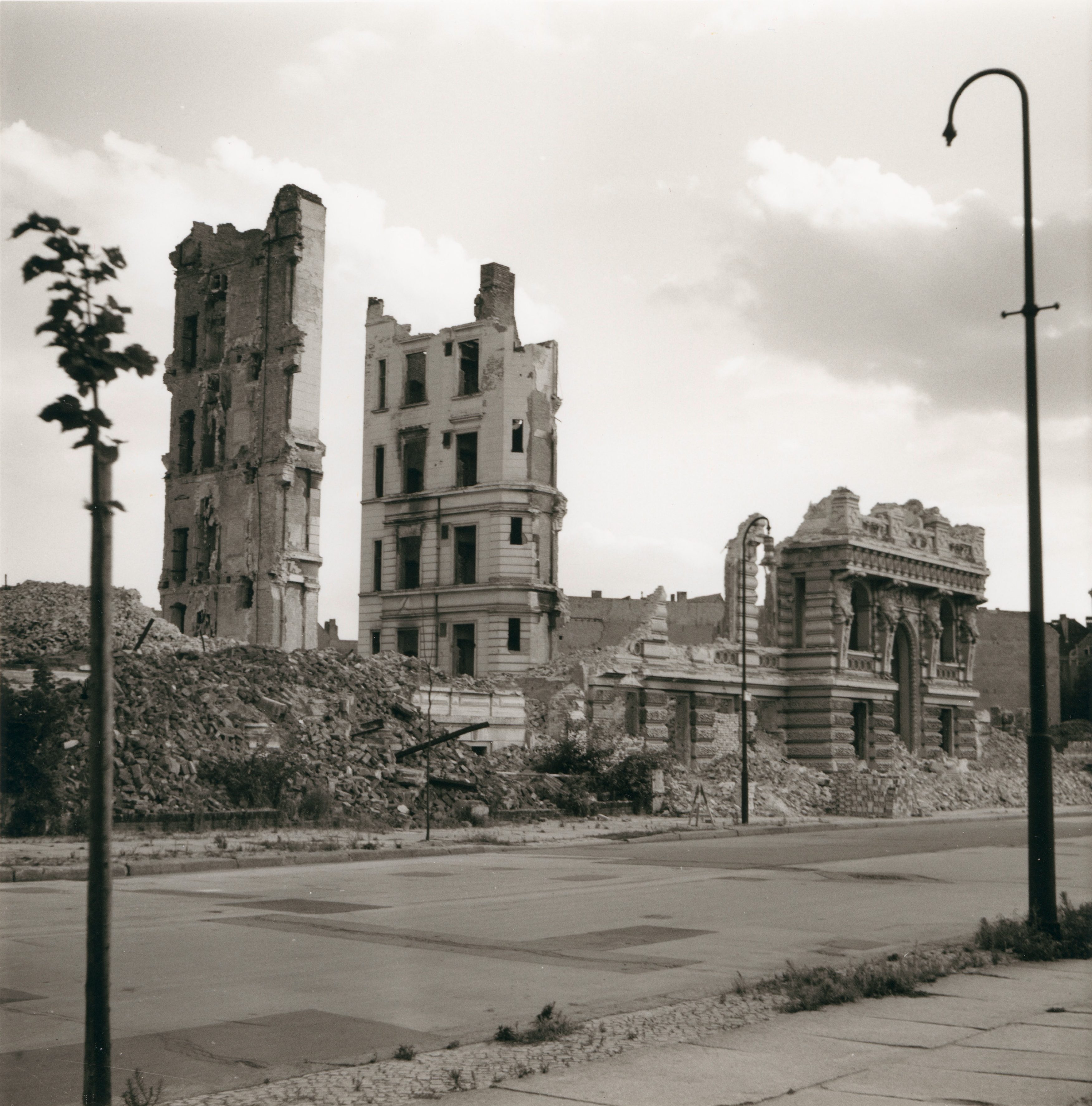 Ruinen von Gebäuden in einer zerstörten Stadtlandschaft, umgeben von Trümmern und einer verlassenen Straße.