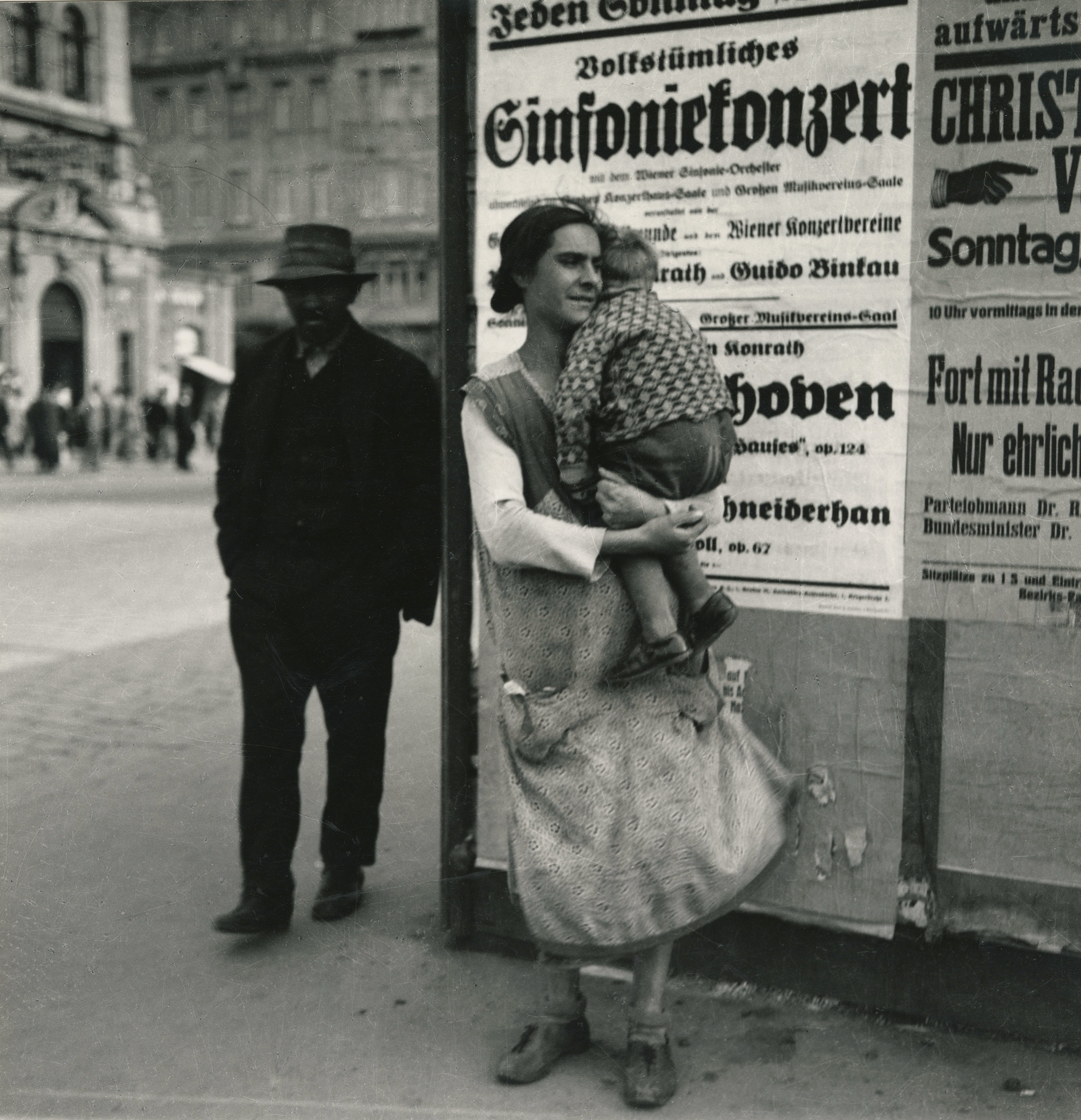 Woman holding a child in front of a billboard, with a man in the background and a historic cityscape in black and white.