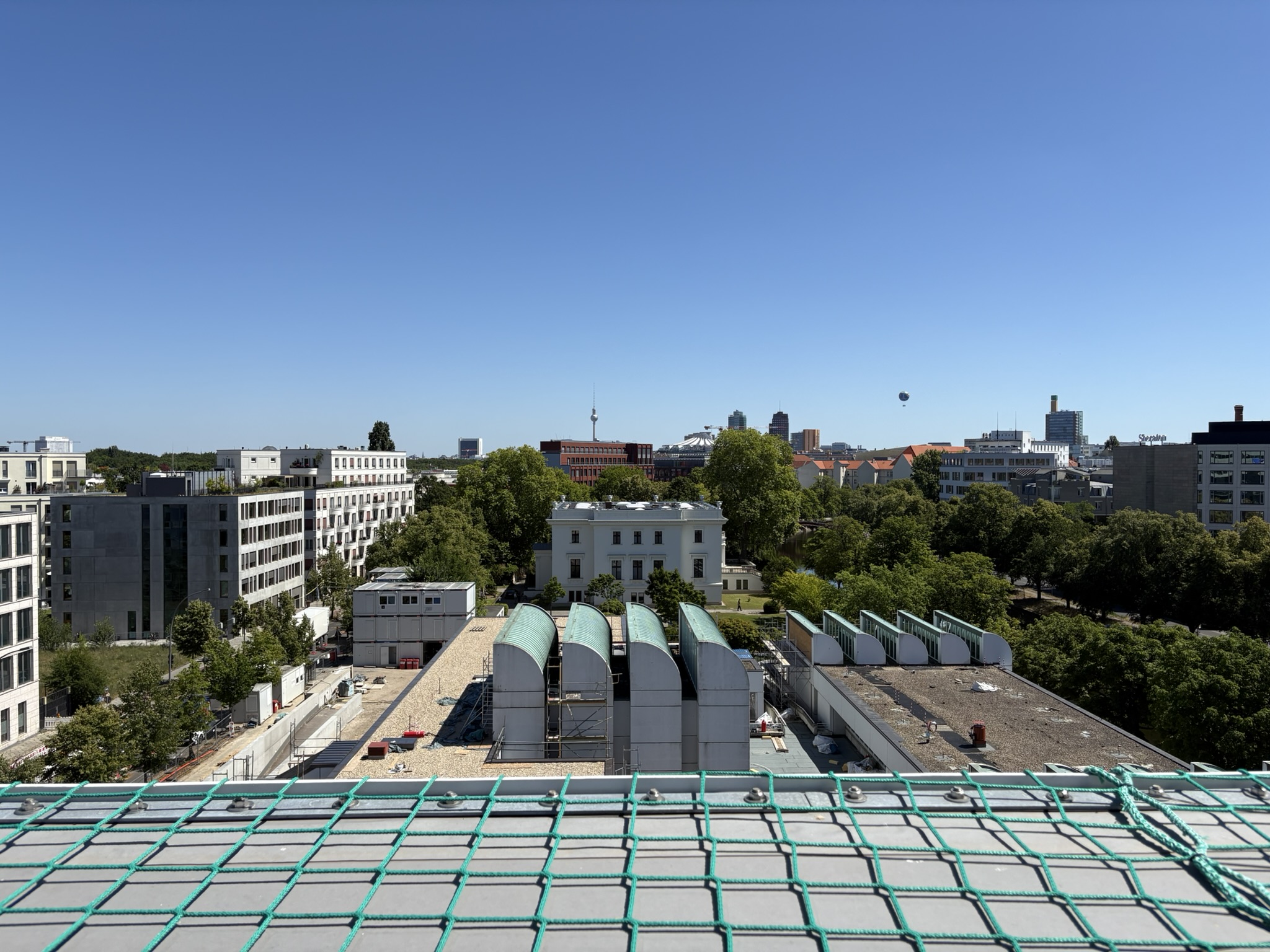 Stadtansicht mit moderner Architektur, Bäumen und einem Ballon unter blauem Himmel. Im Hintergrund der Fernsehturm sichtbar.