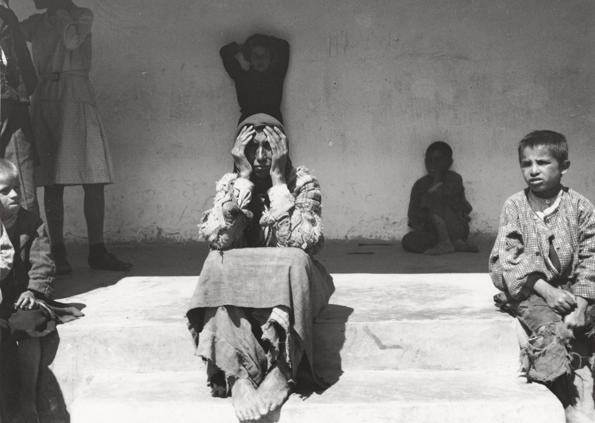 Eine traurige Frau sitzt auf Stufen, umgeben von Kindern, während sie sich die Hände vors Gesicht hält.