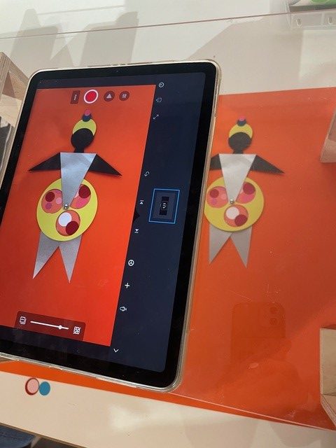 Ein Tablet liegt auf einer Glasplatte. Auf dem Tablet sieht man die Oberfläche einer Stop-Motion-App, die die gebastelte Papierfigur auf orangefarbigem Grund wiedergibt, die darunter auf dem Tisch liegt.