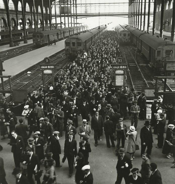 Menschenmenge am Bahnhof, historische Züge und geschäftiger Reiseverkehr zwischen den Gleisen.