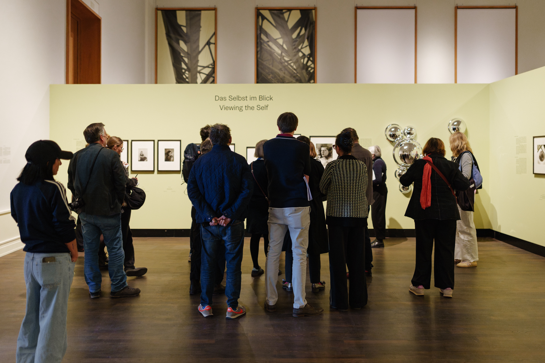Besucher betrachten Fotografien und Kunstwerke in einer Galerie mit grünem Wandhintergrund und Spiegelobjekten.