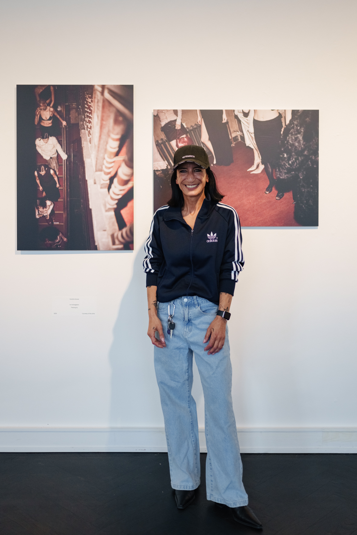 Frau in lässiger Kleidung mit Cap und Sneakers lächelt vor zwei Bildern in einer Galerie.