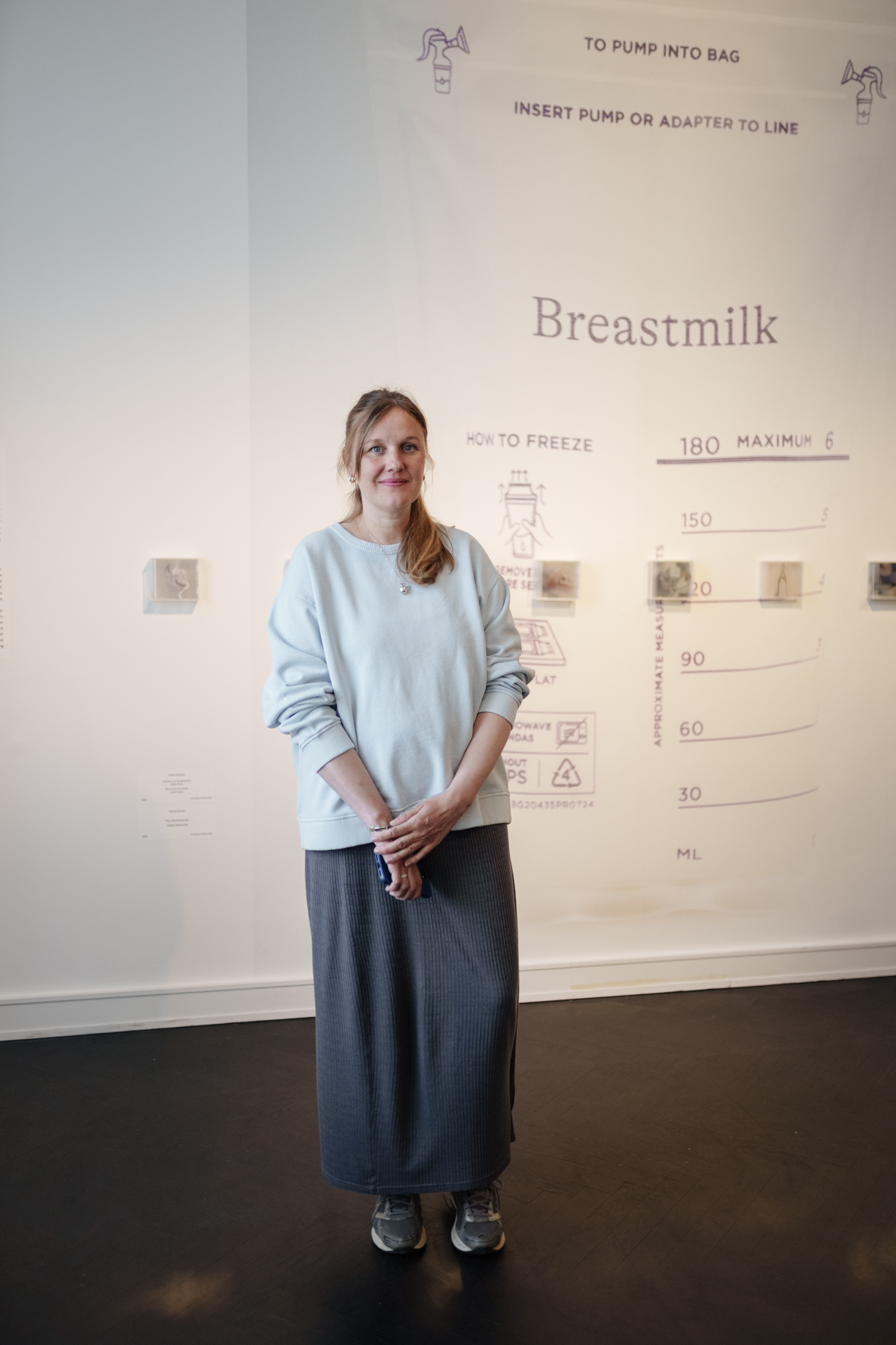 Frau in beiger Oberbekleidung und grauem Rock vor einer Wand mit Informationen zum Milchabpumpen.