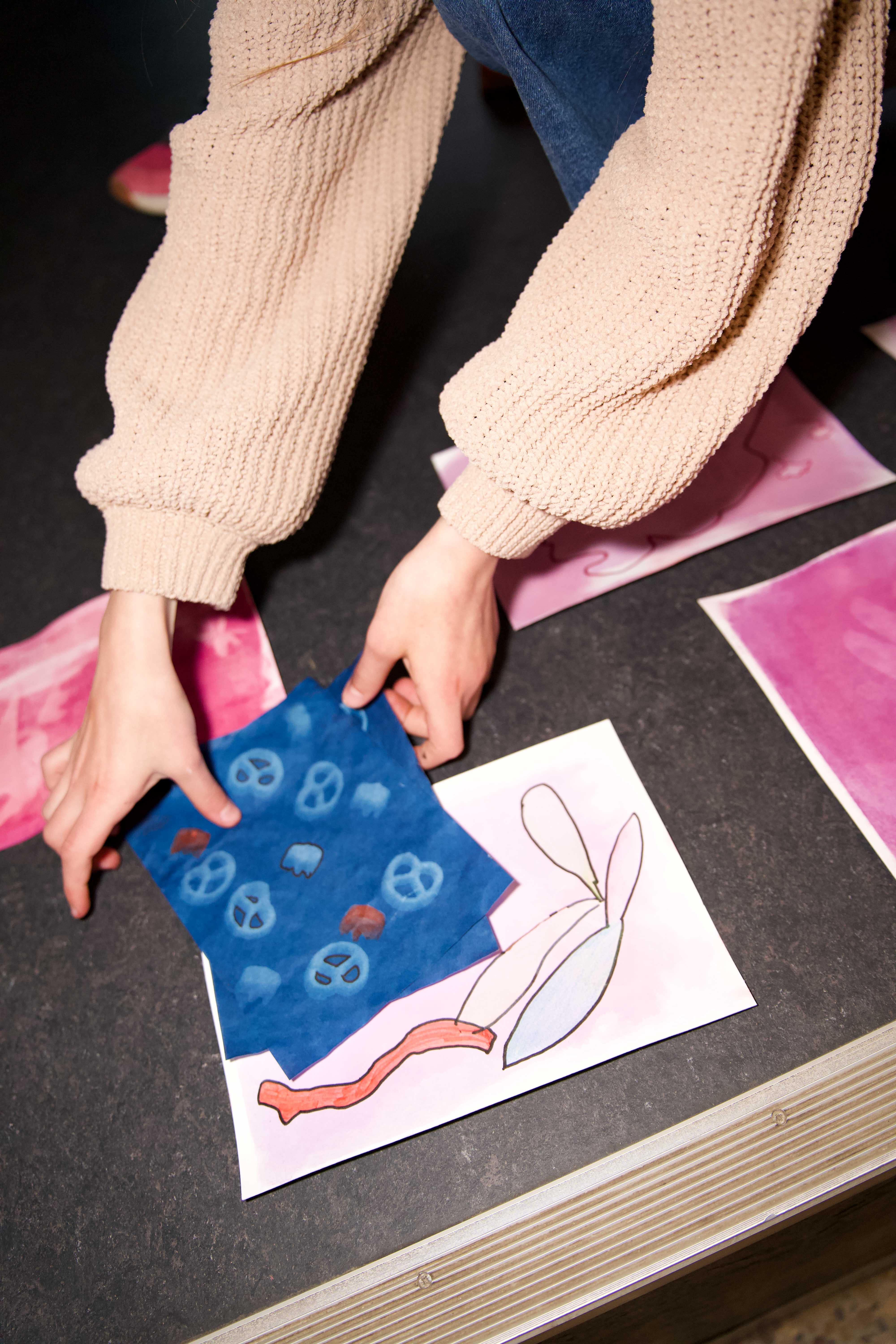 Hände einer Person platzieren farbige Zeichnungen auf einem Tisch, darunter verschiedene Aquarellkunstwerke.