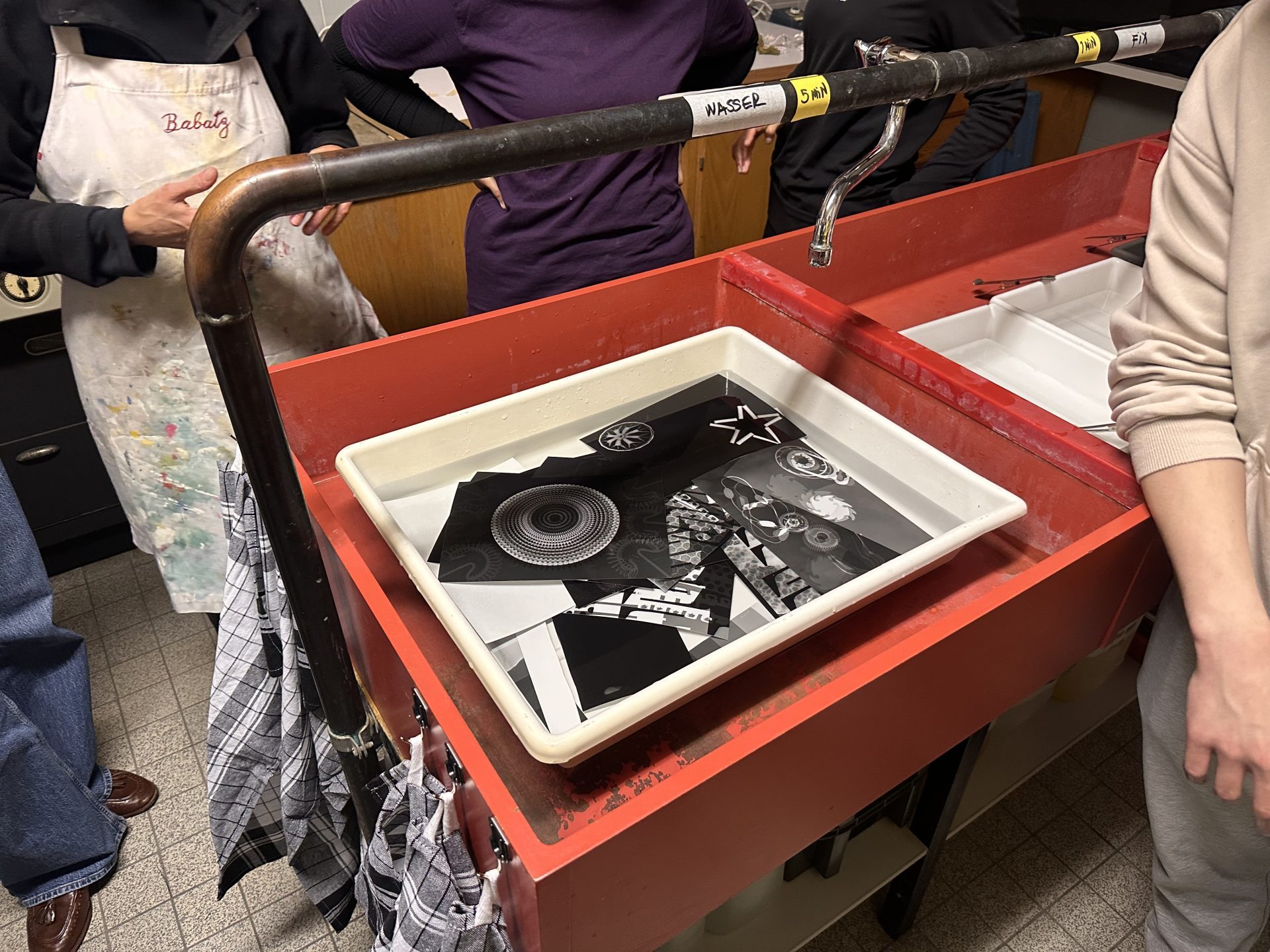 Black and white photo prints in a water tub, surrounded by people in a creative environment.