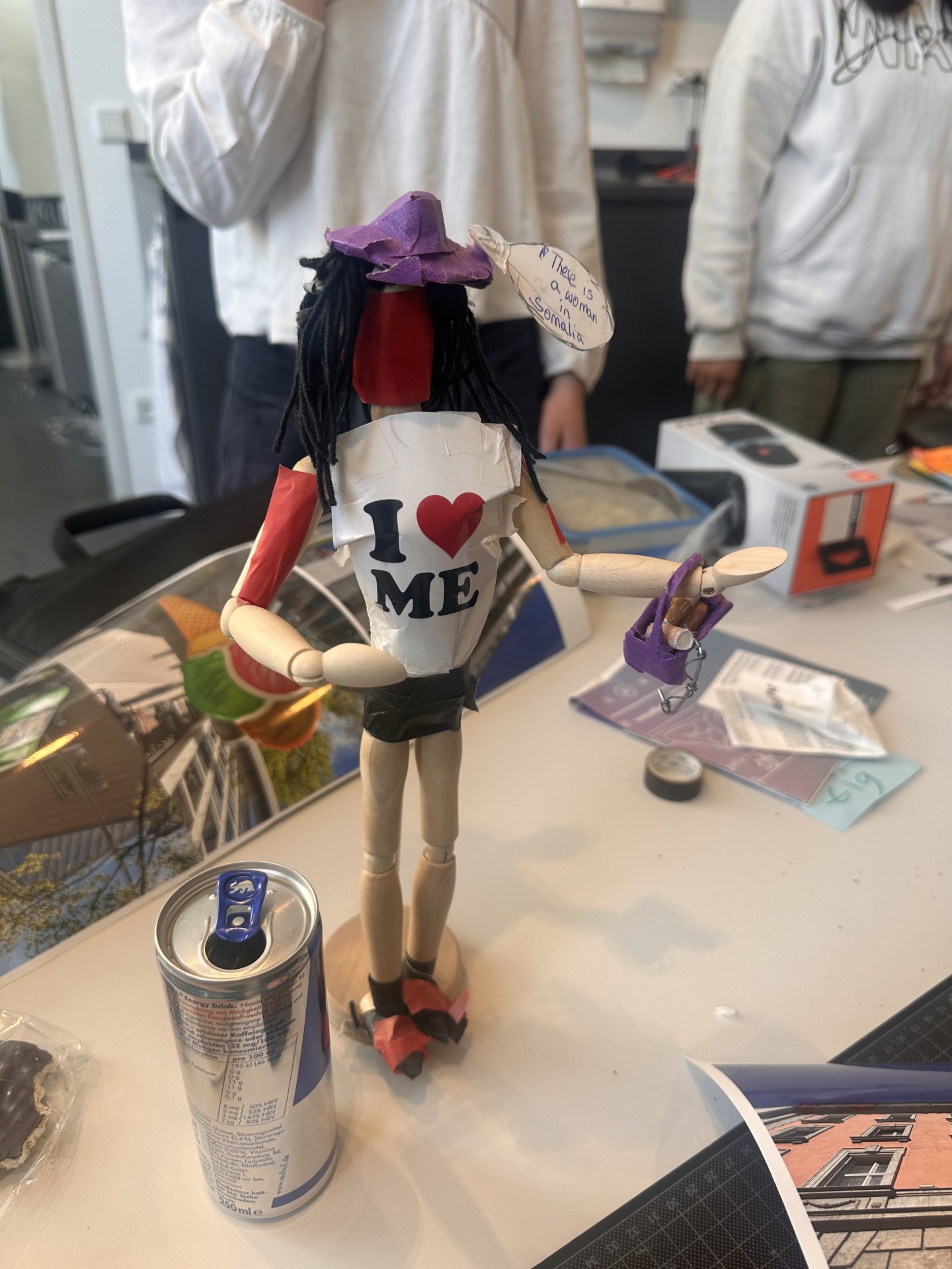 A handmade doll with an "I ❤️ ME" T-shirt, purple hat, and dreadlocks stands on a table next to a drink can.