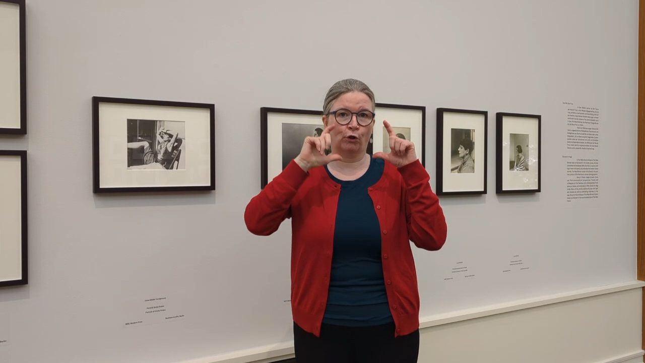 Eine Person mit Brille und rotem Cardigan steht vor Fotos im Museum und erklärt etwas dazu.