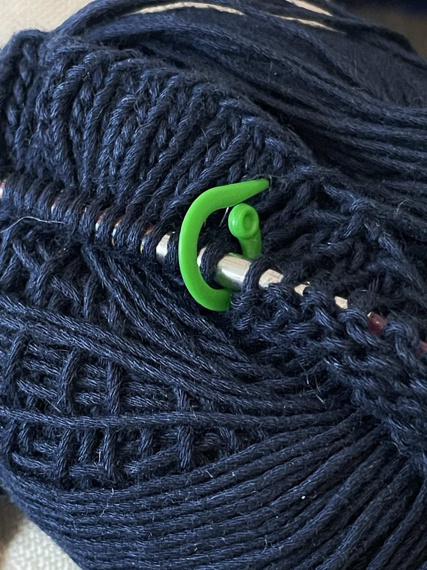 Zu sehen ist ein dunkelblaues Garnknäuel, in dem eine metallene Stricknadel mit aufgezogenen Maschen steckt. Zwischen zwei Maschen befindet sich ein hellgrüner Maschenmarkierer.