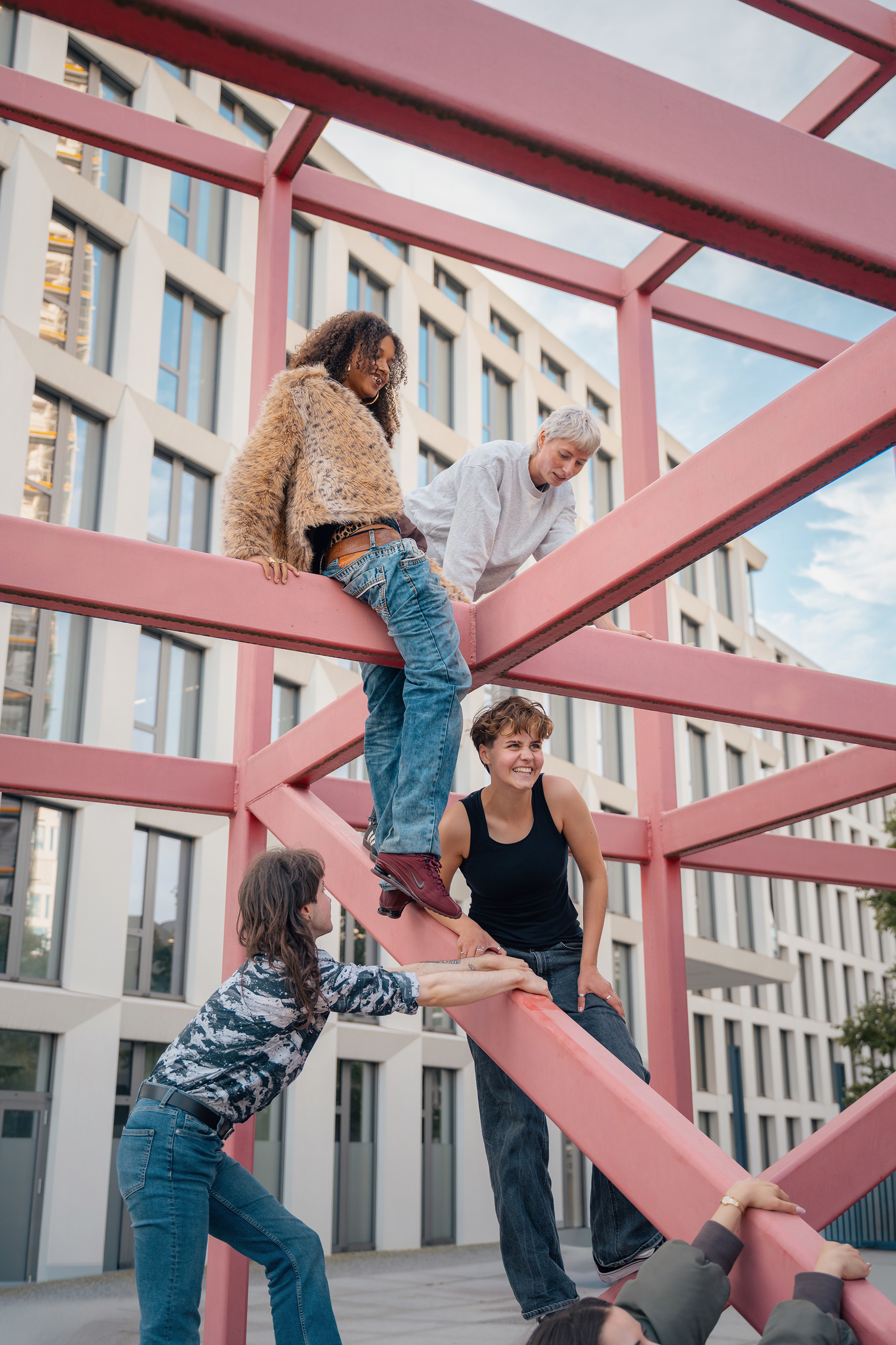 Vier Personen posieren vor modernen Gebäuden im Freien auf einem pinken Metallgestell.