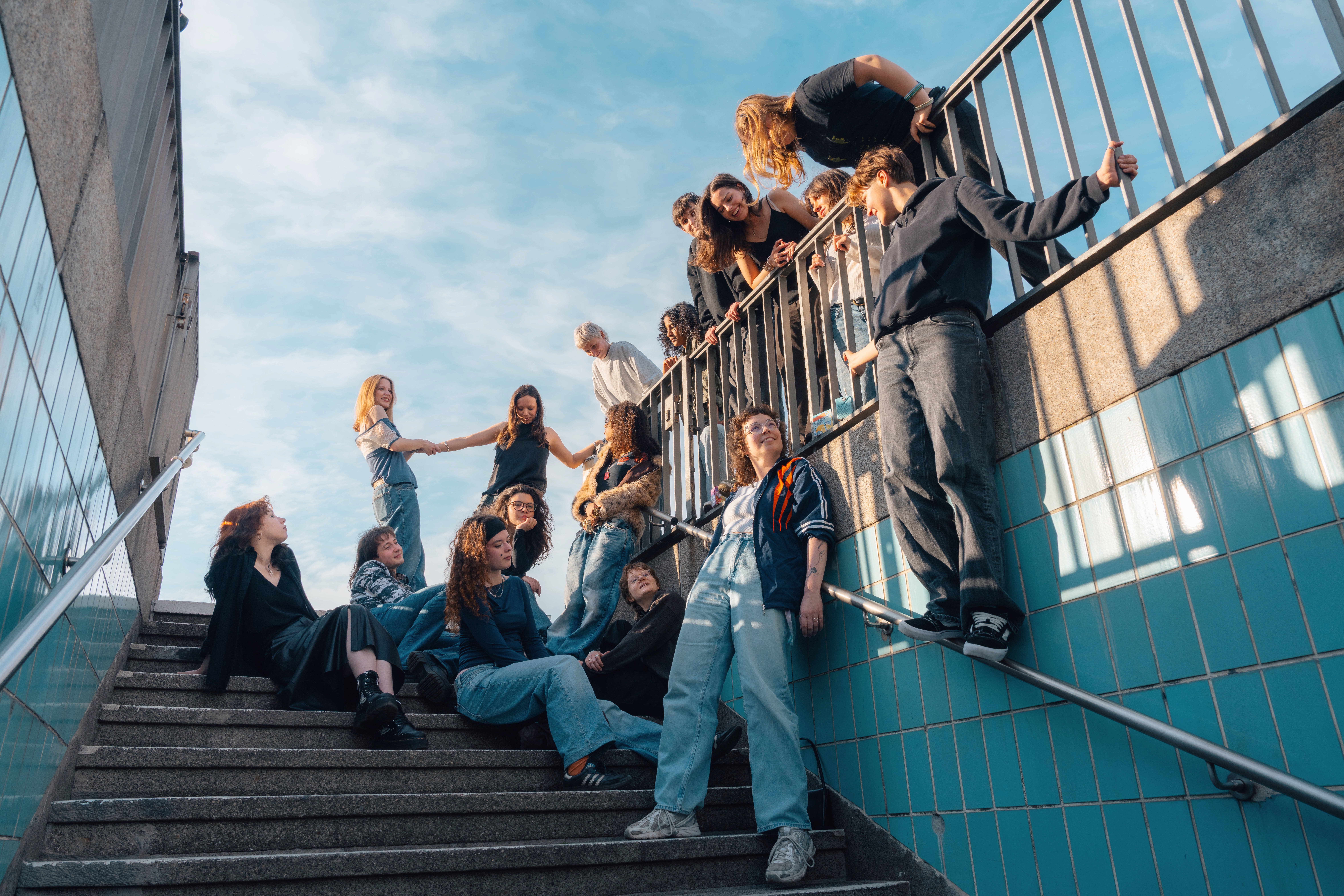 Gruppe junger Leute posiert kreativ auf einer Treppe unter blauem Himmel, in lässigen Outfits und mit fröhlichen Gesichtsausdrücken.