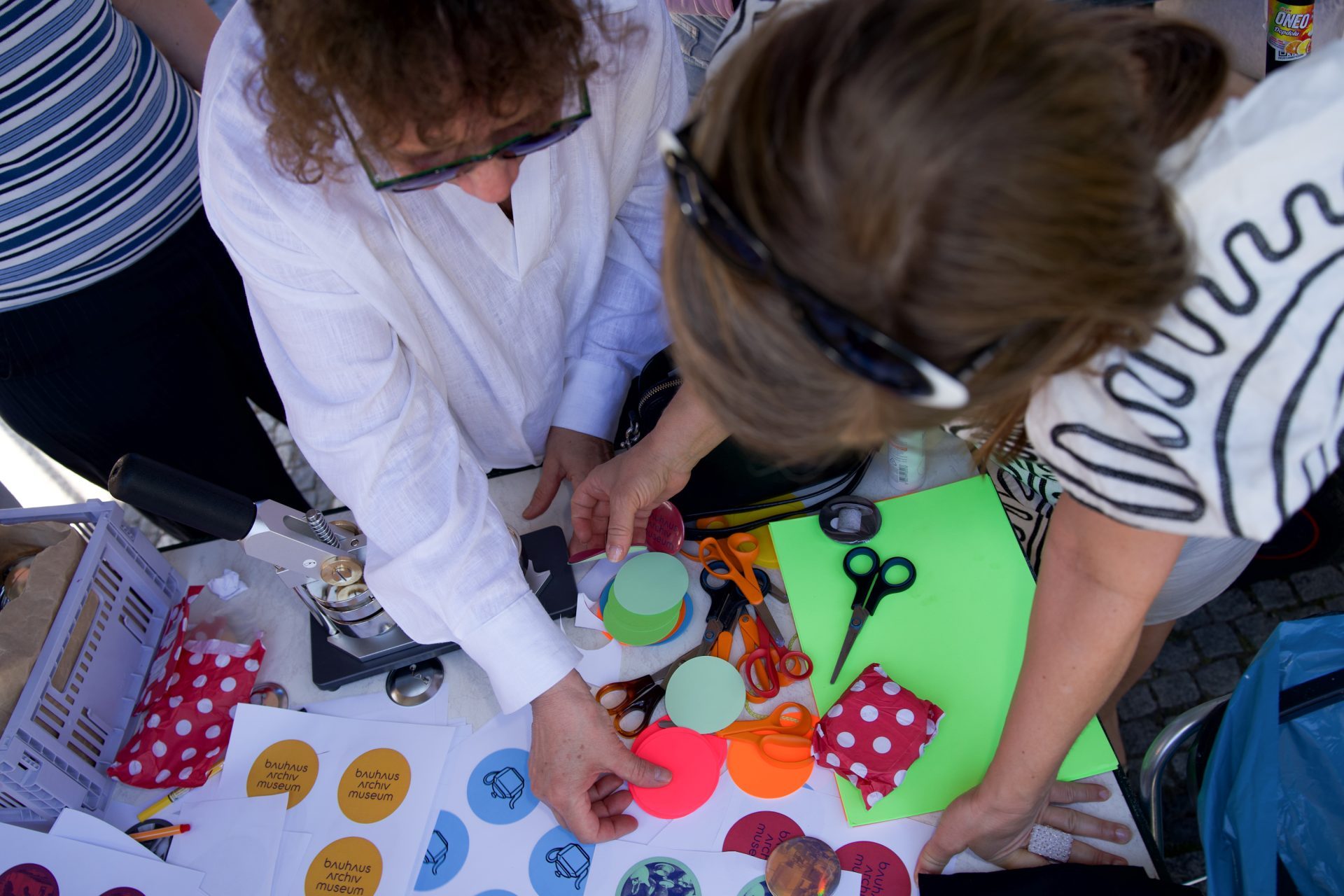 Zwei Personen arbeiten an einem Basteltisch mit Scheren, buntem Papier und einem Button-Maker.