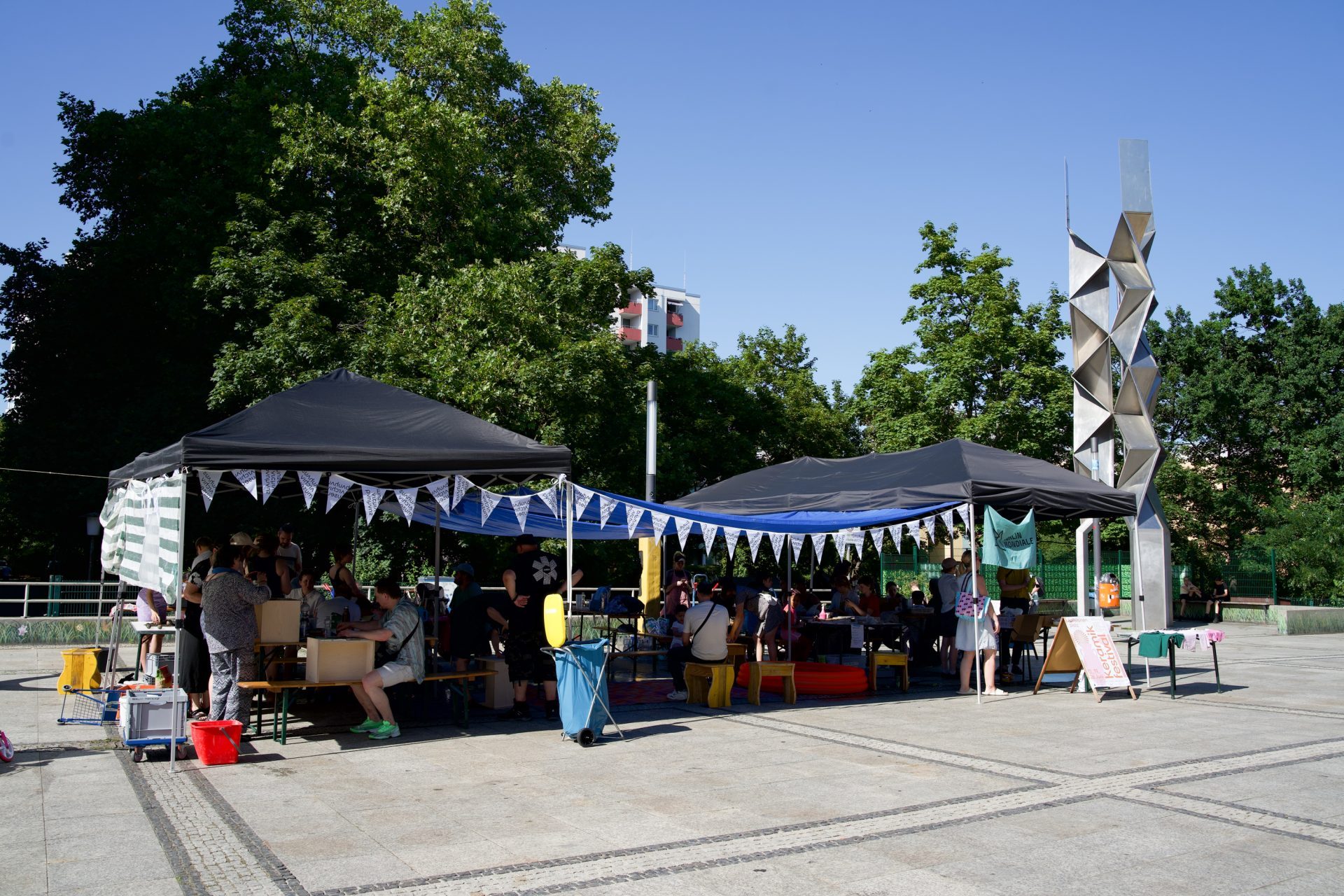 Market with colorful stalls under tents, people interacting, surrounded by trees and a modern sculpture.