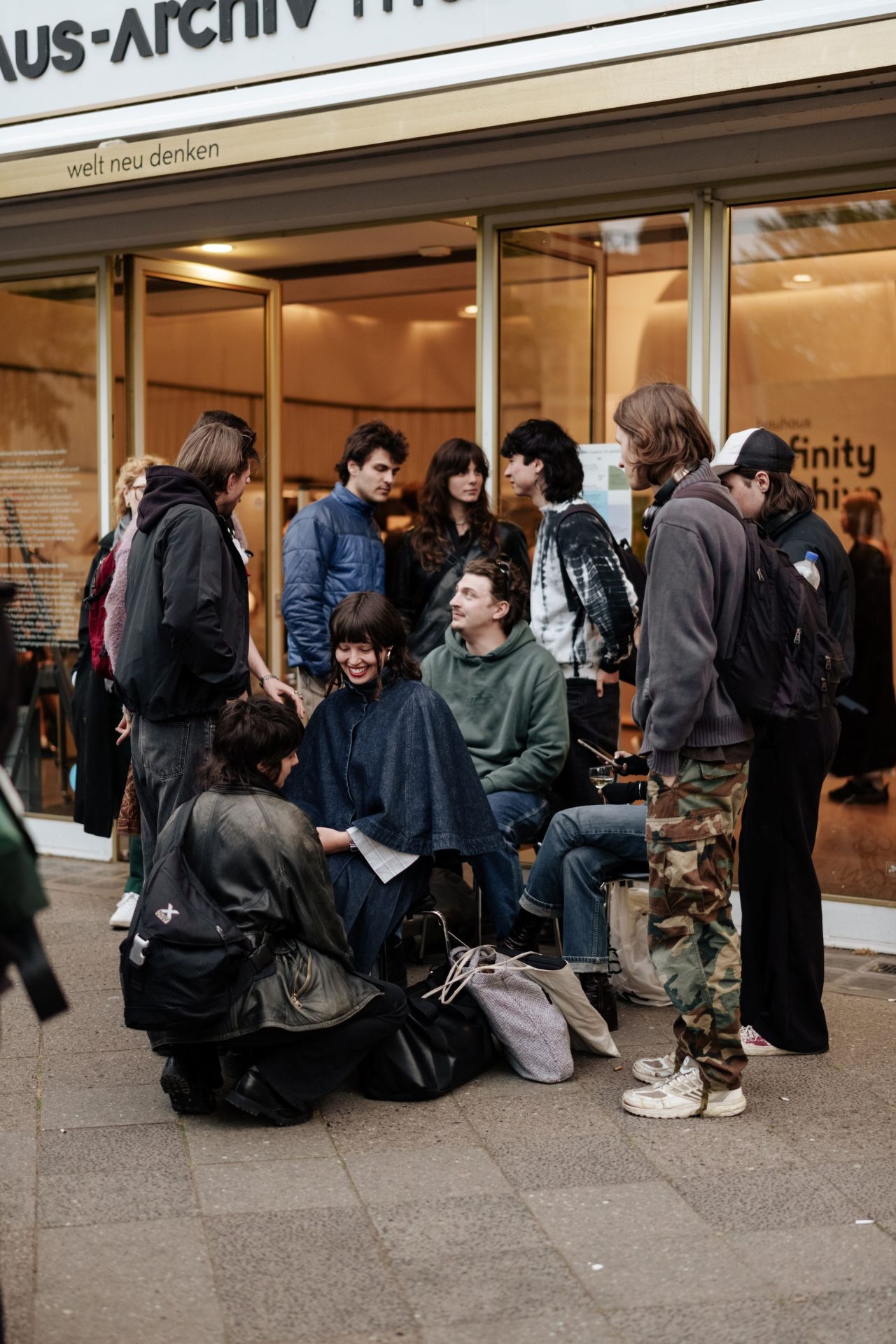 Young people are standing and sitting in front of a shop’s floor-to-ceiling windows.