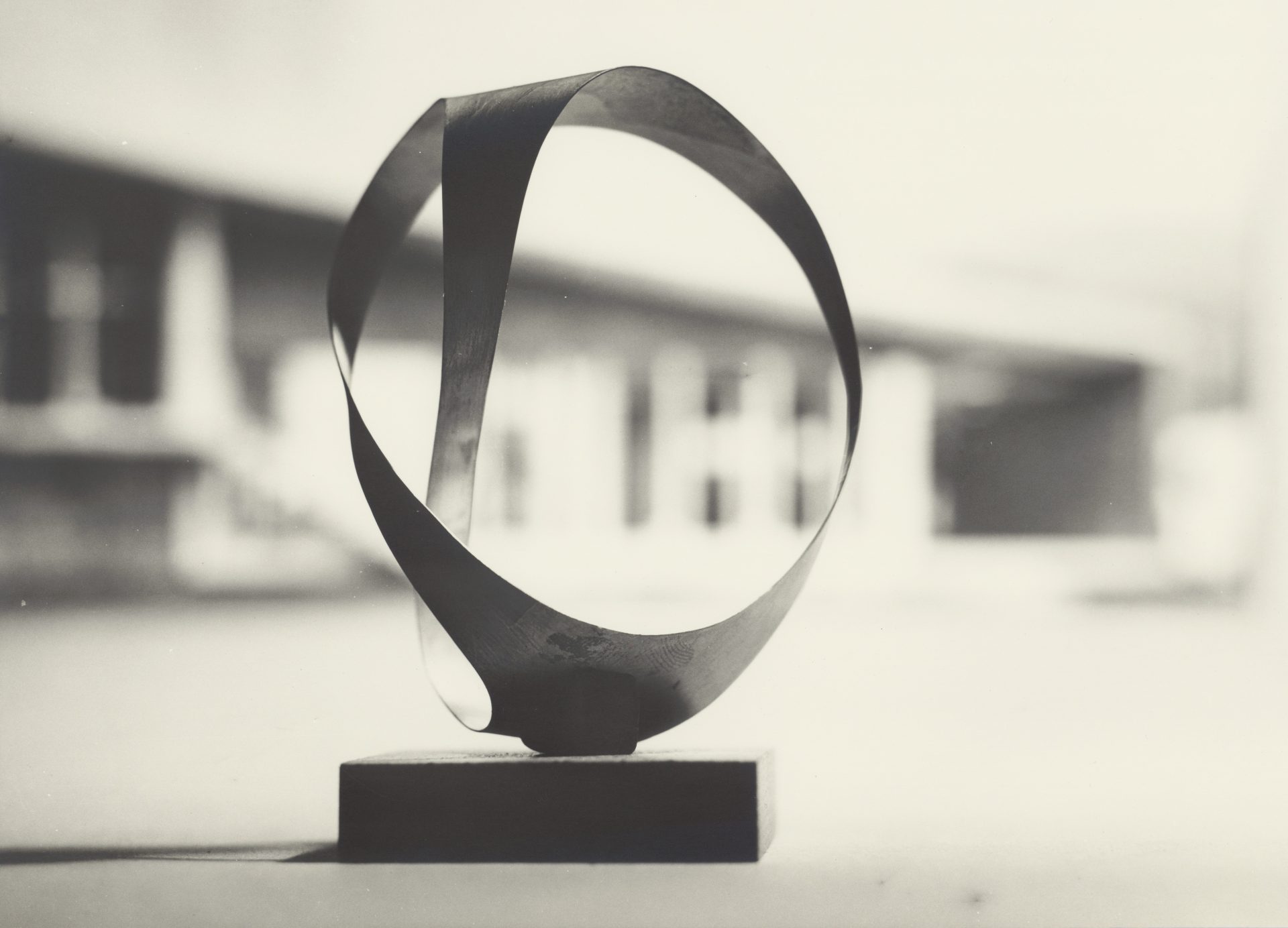 Simple artistic metal object on a wooden base, against a blurred black-and-white background.