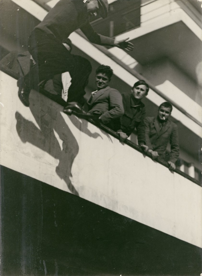 Junge springt von einem Balkon, während andere zuschauen und lachen. Schwarz-Weiß-Foto, nostalgische Atmosphäre.