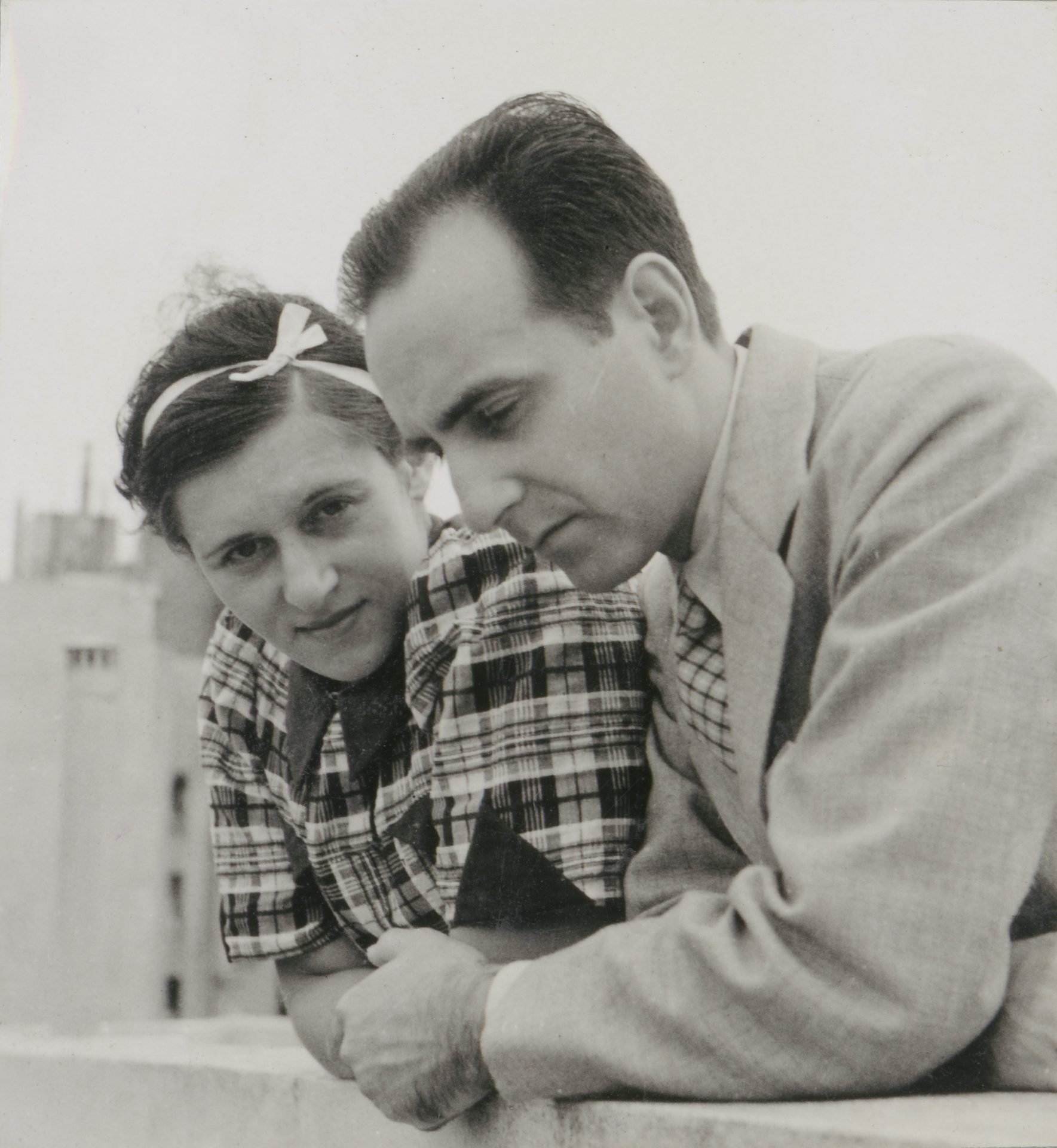 Two people, a woman in a plaid dress and a man in a suit, lean on a railing and look down.