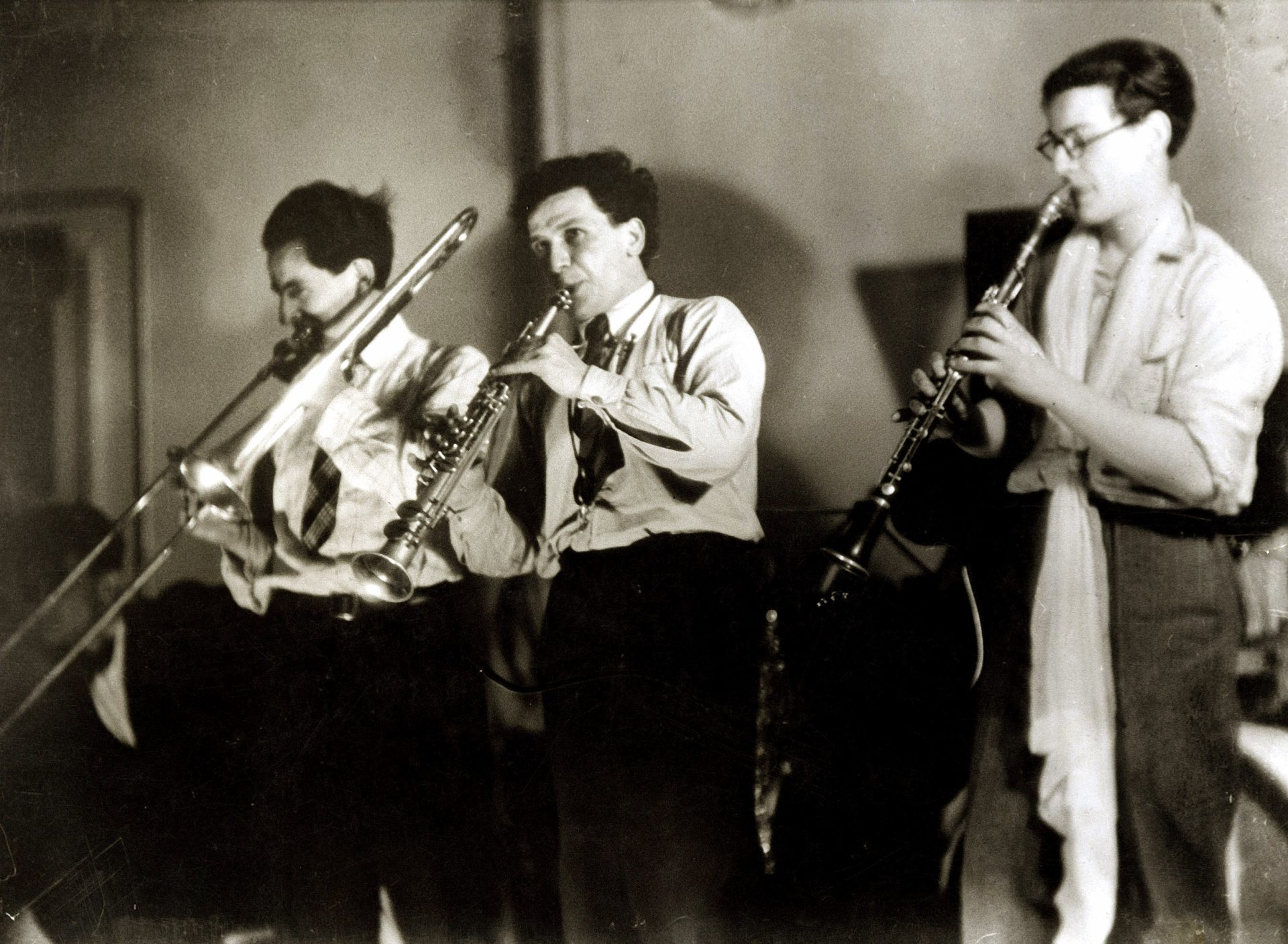 Drei Männer in dunklen Hosen und hellen Hemden geben mit ihren Blasinstrumenten ein Konzert. Der Mann links spielt Posaune, der Mann in der Mitte Klarnette und der Mann rechts Oboe.