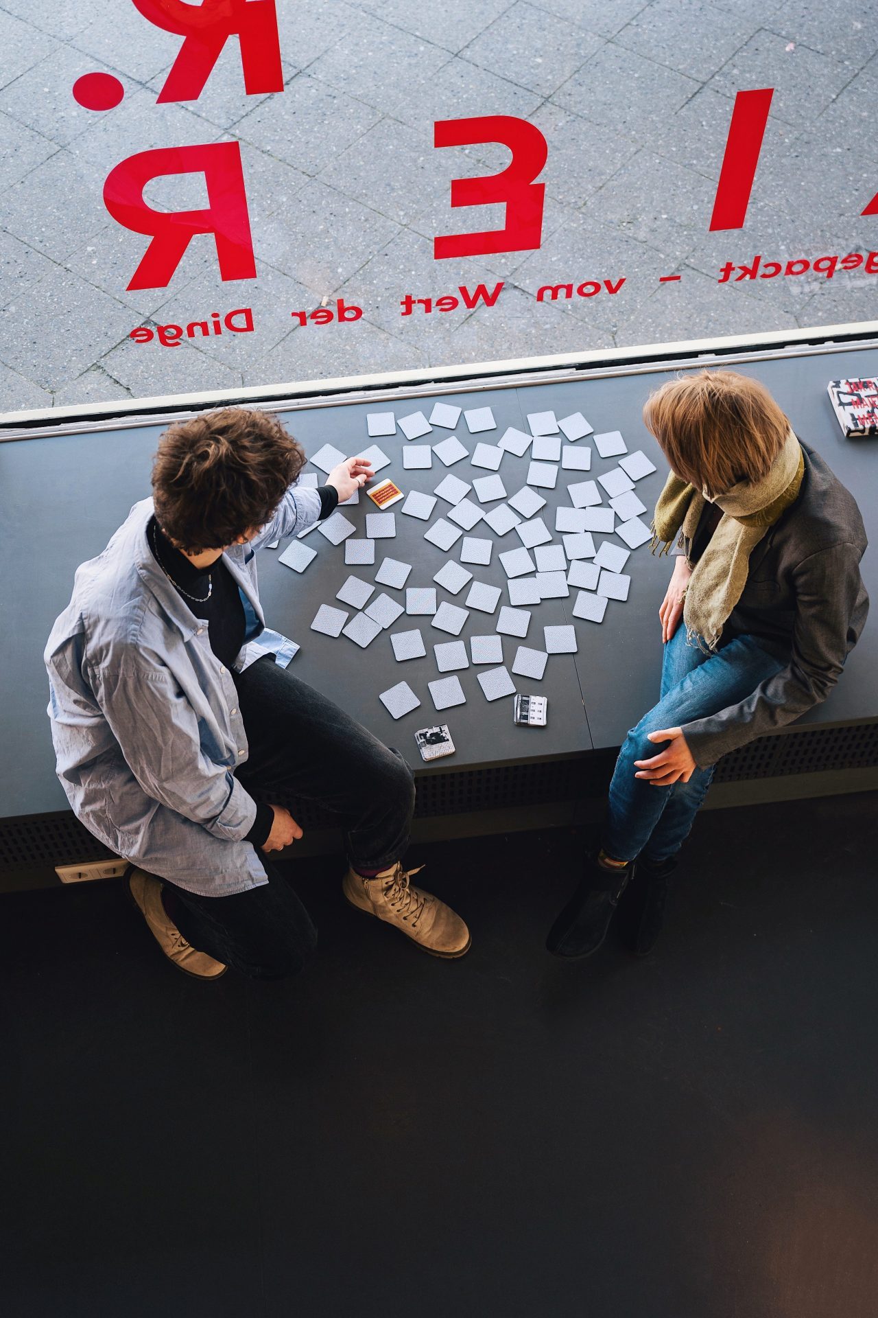 Two people sit at a table playing with numerous cards, surrounded by large labeled windows.