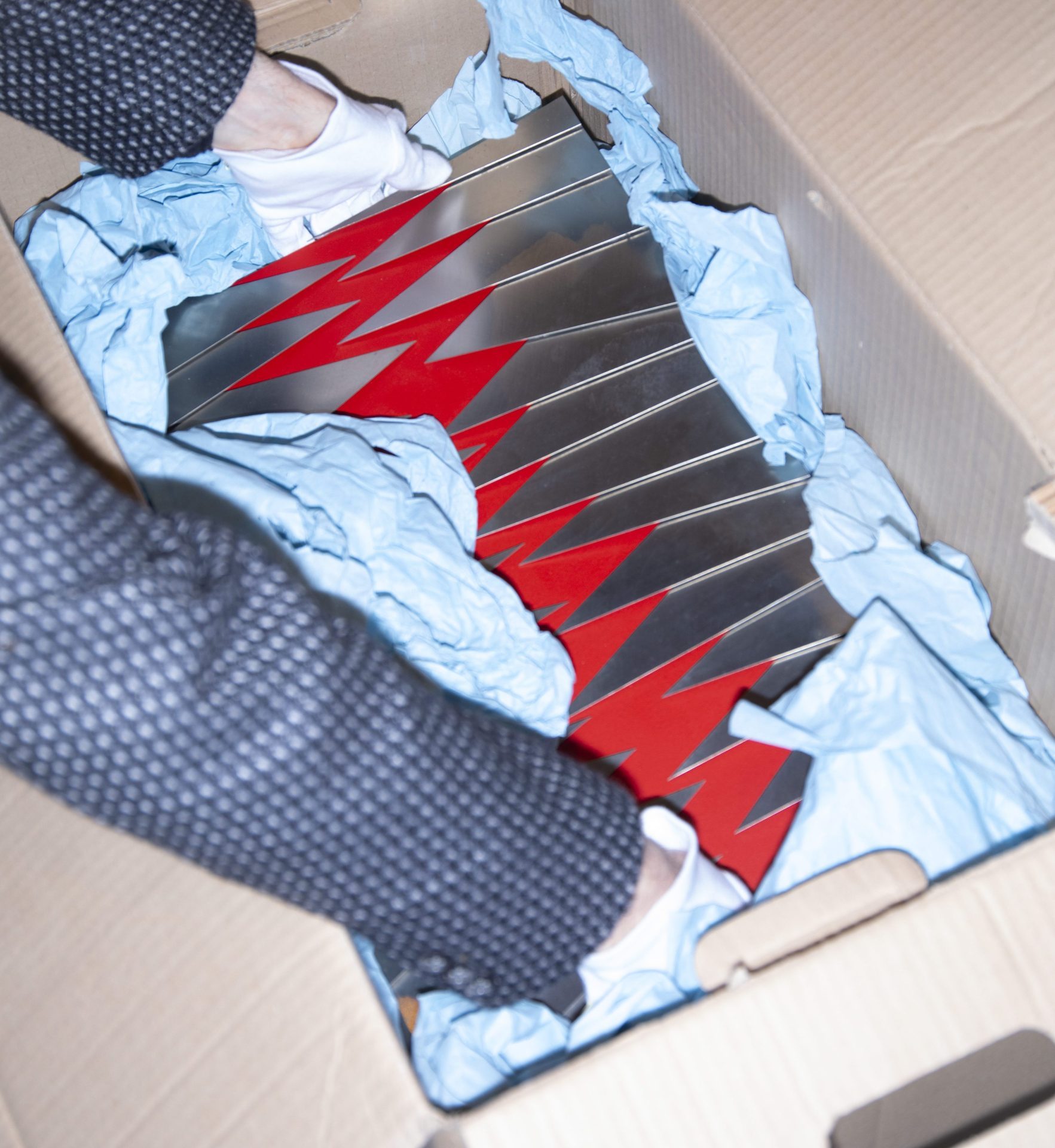 A hand lifts shiny, red-metallic objects from a box, surrounded by blue packaging foam.