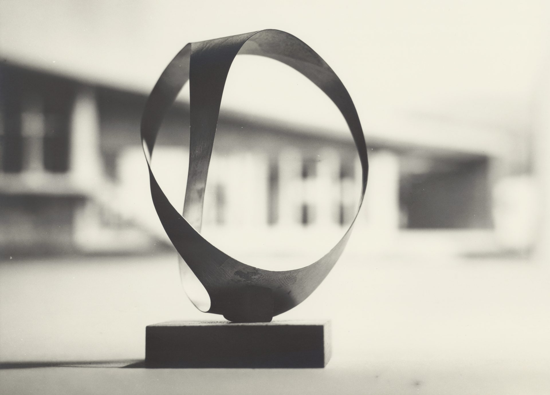 Abstracte Skulptur aus Metall, die eine fließende Form auf einem rechteckigen Sockel darstellt. Schwarz-weiß Fotografie.