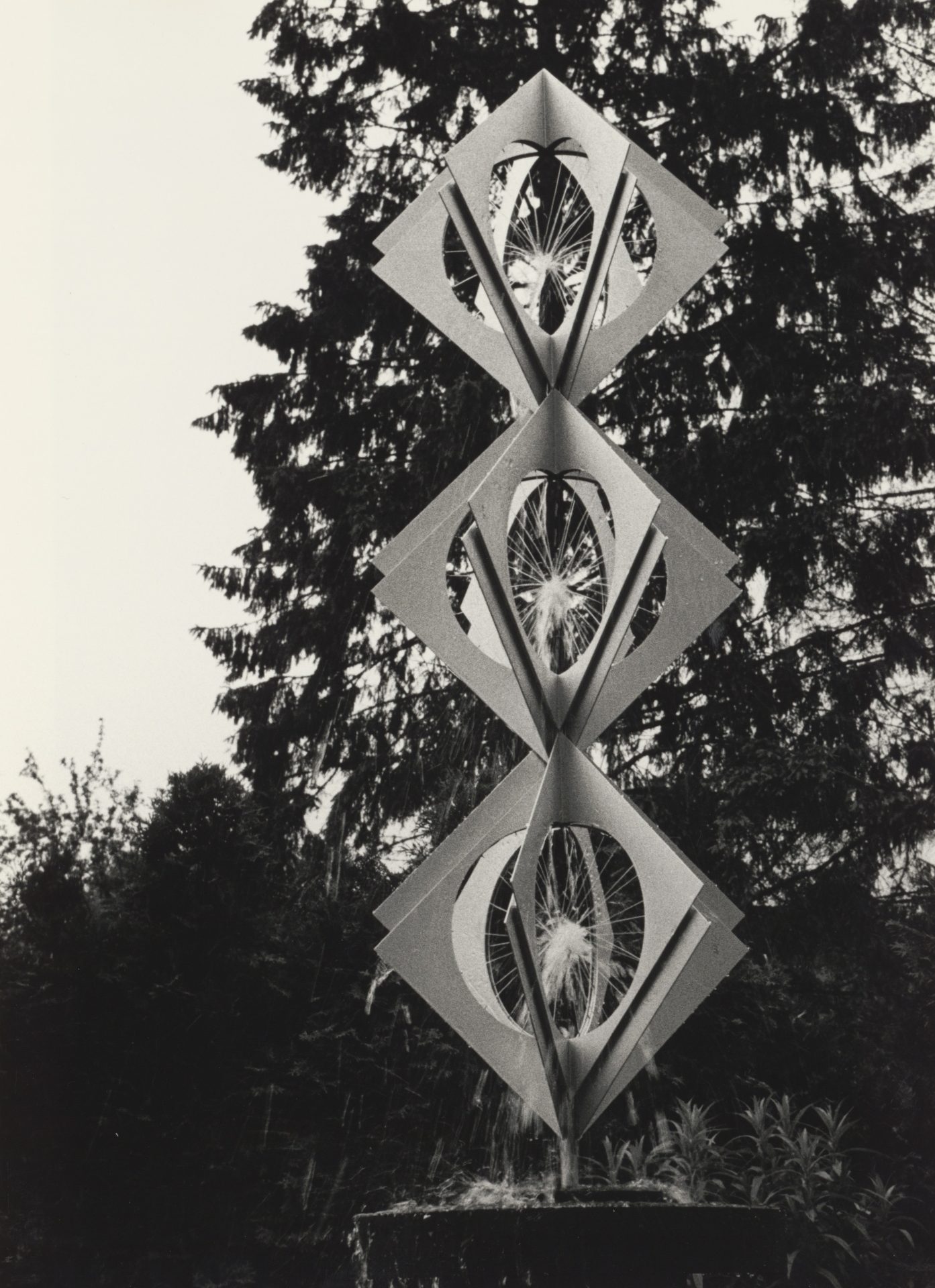 Sculpture made of paper and metal with geometric shapes, surrounded by trees, in a black and white photo.