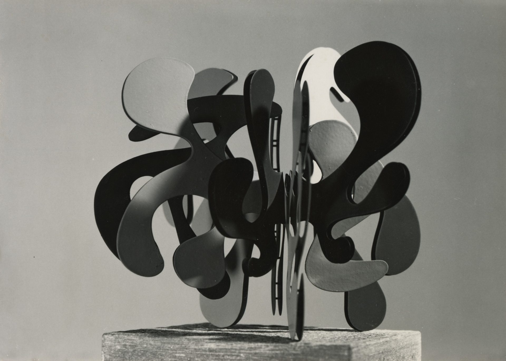 Abstract sculpture made of curved black and gray shapes on a wooden pedestal, against a neutral background.