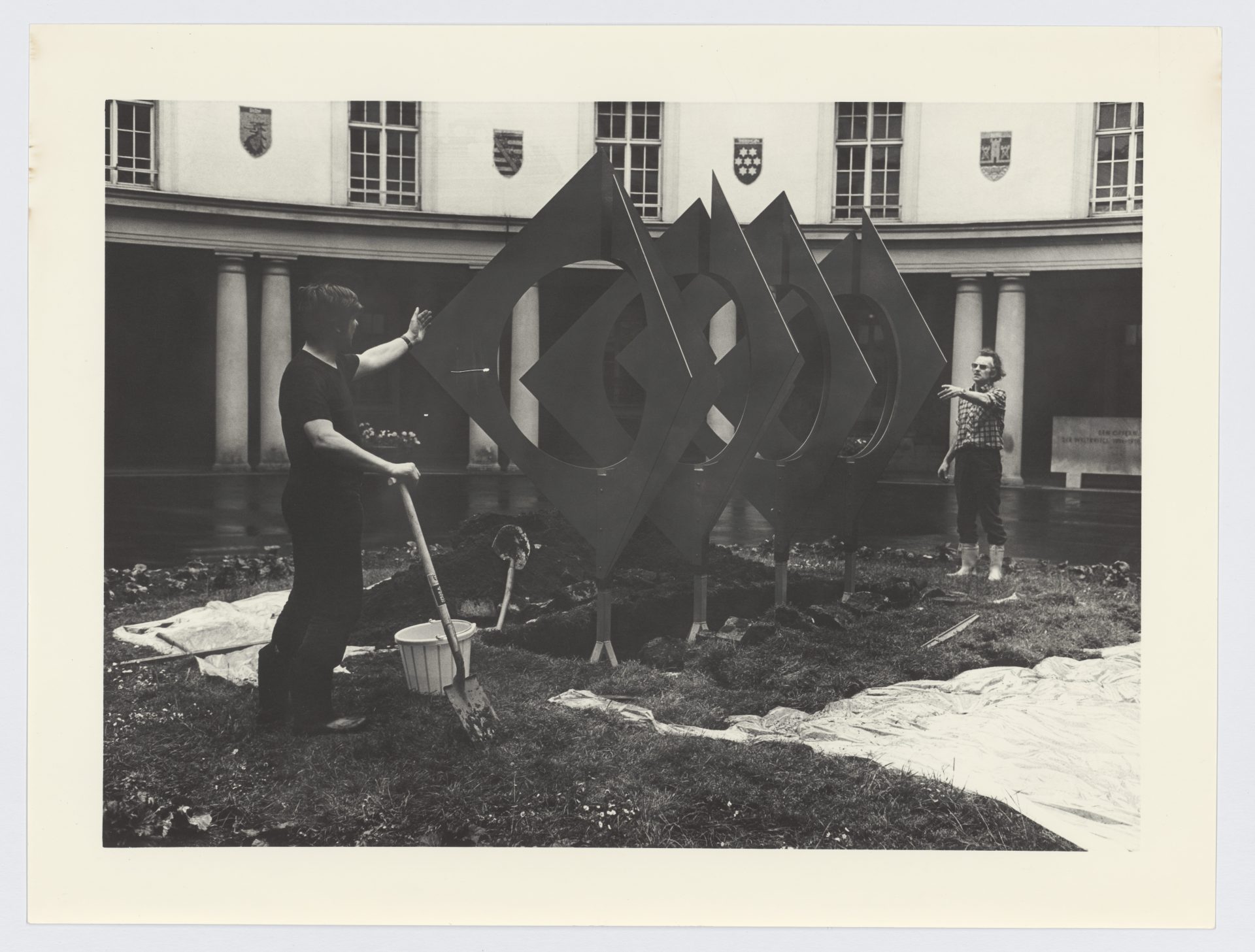 Two people are working on an outdoor art installation surrounded by sculptures and a wet ground.