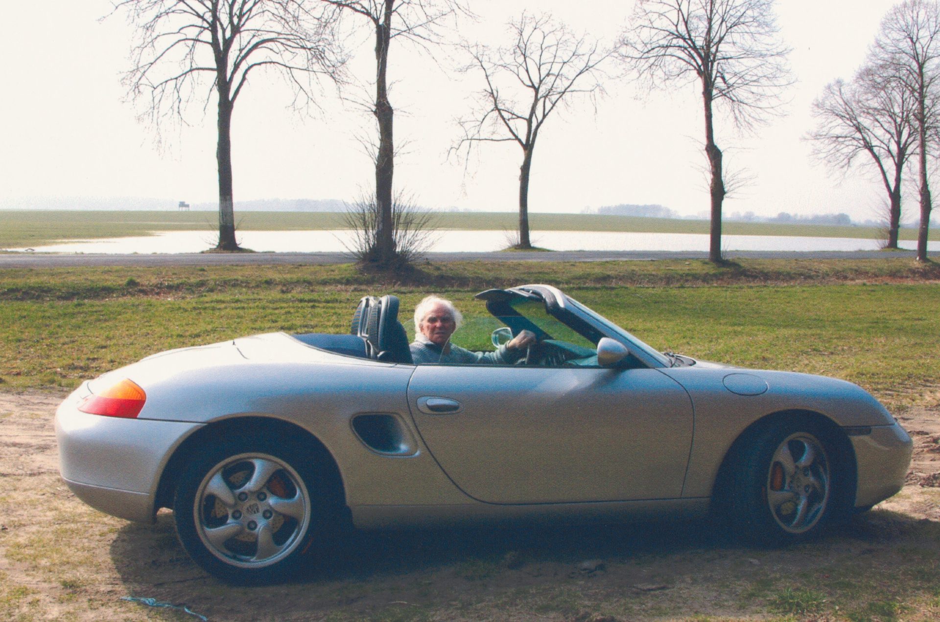 An older man is smiling in a silver convertible, sitting on a meadow with trees and water in the background.