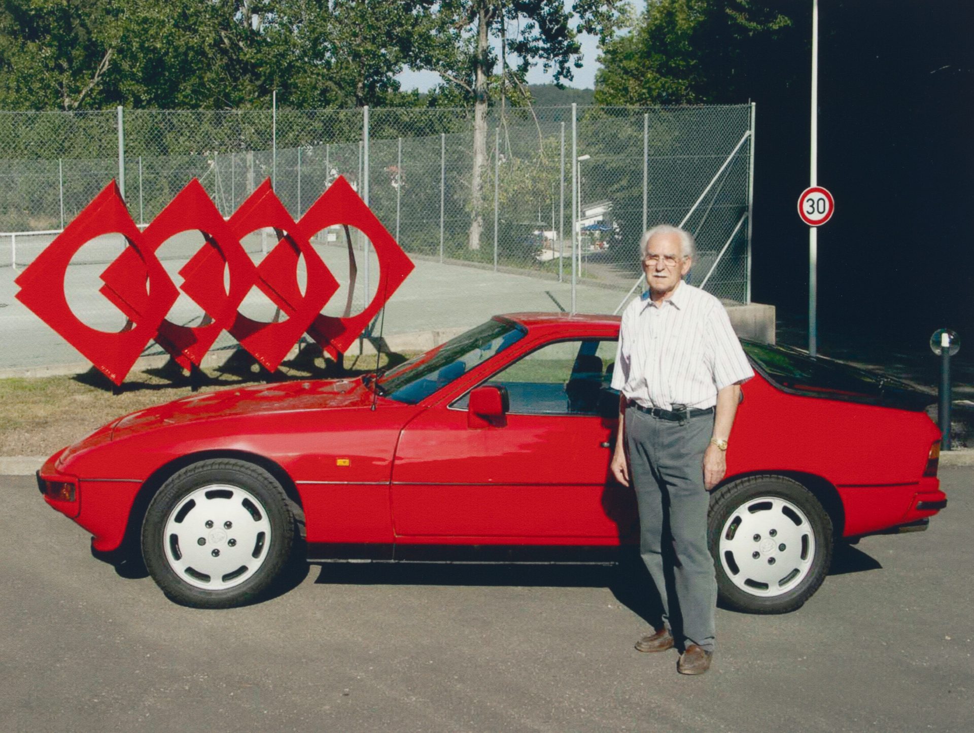 An elderly man stands next to a red car in front of a sculptural installation made of red diamonds.
