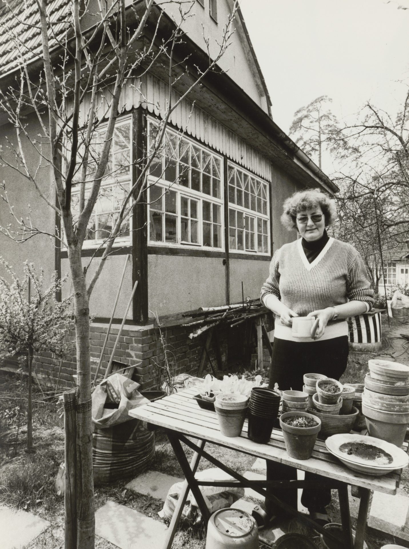A woman with glasses and curly hair stands at a table with plant pots in front of a house in the garden.
