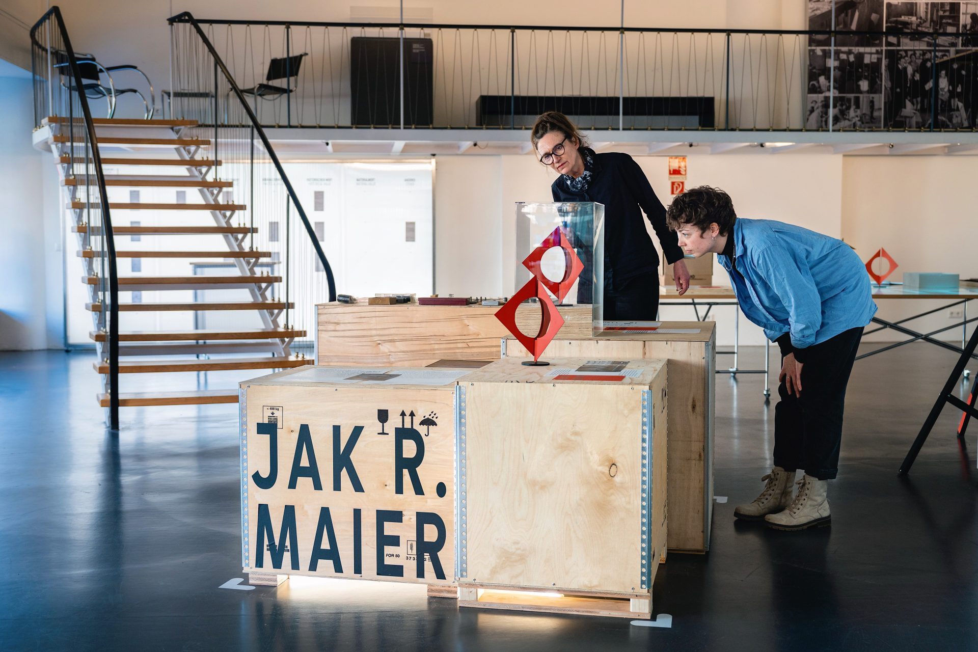 Two people look at art objects in a modern exhibition hall with wooden sculptures and a staircase in the background.