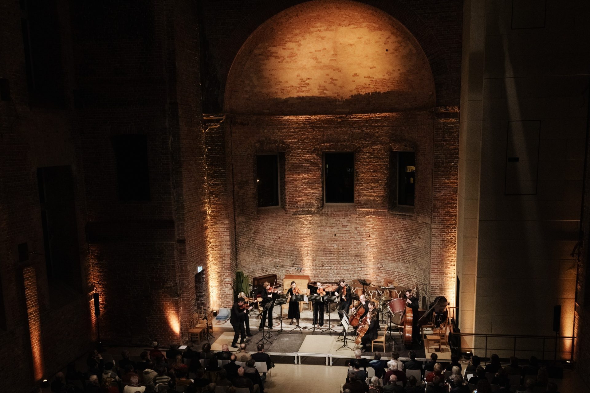 An orchestra plays in a historic hall softly illuminated, in front of rows filled with audience members.